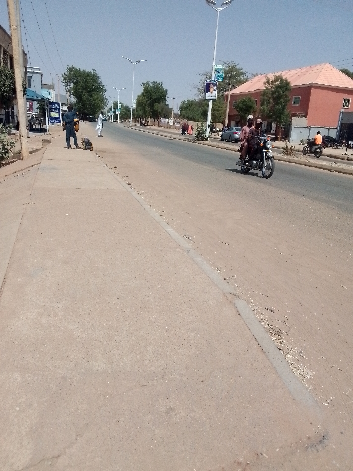 A sunlit street scene shows a two-lane road with a wide, sandy shoulder and a concrete sidewalk. Several people are visible, including two men on a motorcycle, a man standing on the sidewalk, and a few others in the distance. Buildings, trees, and streetlights line the road. A campaign poster can be seen on a pole. The sky is clear.