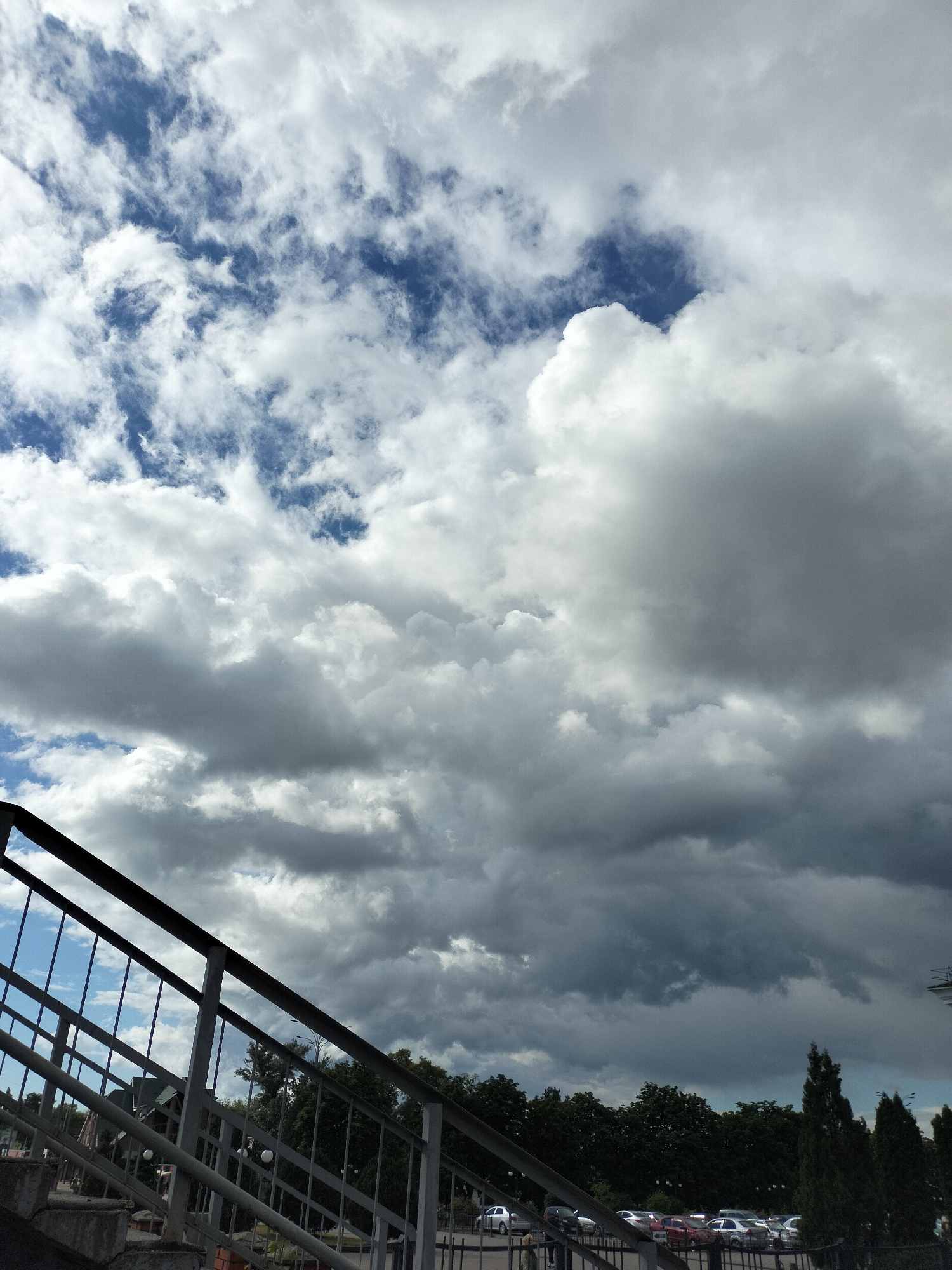 The image captures a scene with a view dominated by a dramatic sky filled with a mix of white and grey clouds. Patches of blue sky are visible through the clouds. A metal staircase with railings is visible in the foreground. The staircase overlooks a tree line and a parking lot with several cars. The location appears to be in an urban area within the city of Unknown, Unknown, likely on an elevated pedestrian walkway or bridge. No people are visible in the shot.