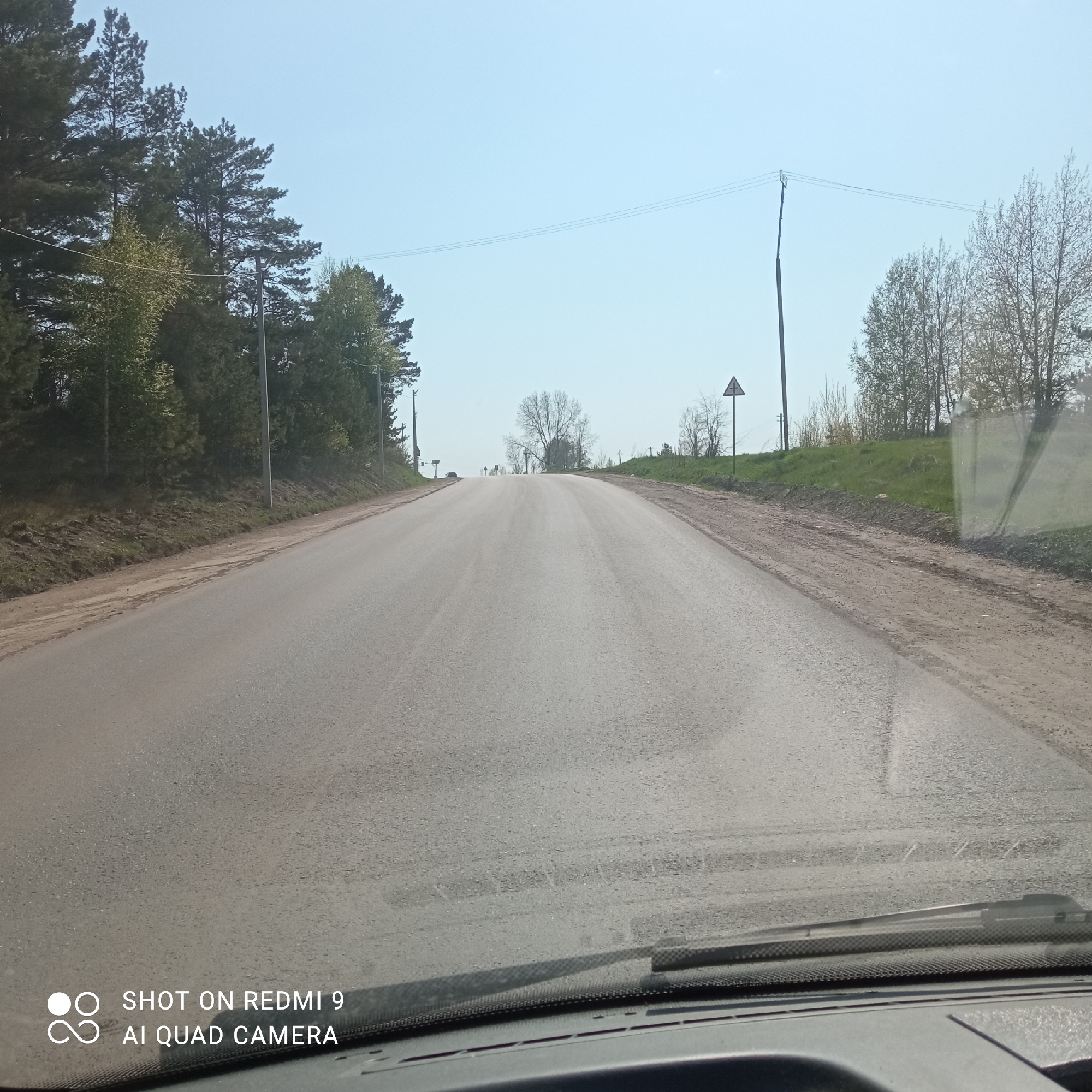 The image depicts a view from inside a vehicle traveling on an asphalt road. The road leads uphill and into the distance, with trees lining the left side. A power line runs parallel to the road on the right. A triangular road sign is visible on the right side of the road. The foreground shows a portion of the vehicle's dashboard and a windshield. The sky is clear. Based on the landscape and infrastructure, the location is likely in a rural or suburban area. However, there are no identifiable landmarks or signs to determine the specific location in Chaikovsky, Russia. More information is needed to confirm the location in this city.