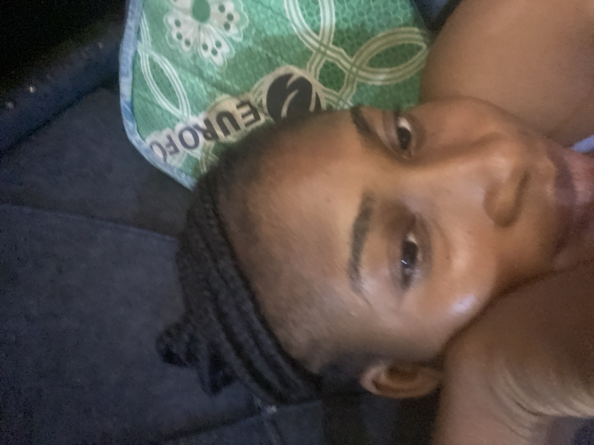 A close-up, overhead shot captures a person lying down, with their head resting on a green pillow. The pillow features a repeating pattern of white floral-like shapes and what appears to be the text "EUROFO" in black lettering. The person's face is partially visible, turned to the left. They have braids in their hair, which is tied back. The lighting is dim, and the background is dark, suggesting an indoor setting, possibly a bedroom. The scene is static, with no visible action or interaction. The image is taken in the city of Kubwa, Nigeria.