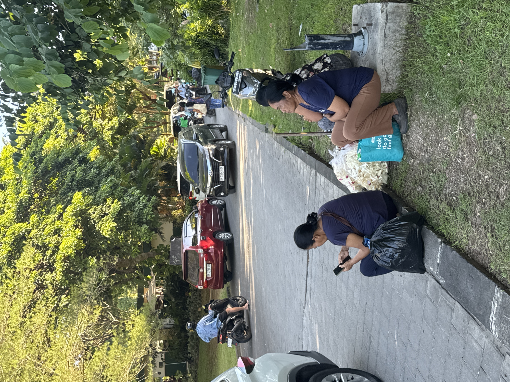 Here's a detailed description of the scene:
**Location:** The image appears to be taken on a paved road or pathway in Kuta Selatan, Indonesia. The presence of lush tropical vegetation suggests a potentially residential or commercial area with planned landscaping.
**People:** Two women are kneeling or crouching alongside the road's edge. One is looking down at a phone, next to a black plastic bag. The other is looking down with her hands held together. Nearby is a motorcycle rider stopped on the road.
**Objects:** Several cars and motorcycles are parked along the roadside. Near the women are a blue shopping bag with words "look, do go, feel g". There are bags that appear to contain white flowers.
**Actions:** The women are engaged in some activity near the roadside, possibly preparing or collecting something. The motorcycle rider is stopped.
**Interactions:** It's unclear if the women are interacting with each other or if the motorcycle rider is interacting with either of them.
**Environment:** The environment includes the paved pathway, a grassy area next to the pathway, and dense tropical trees and foliage. The presence of parked vehicles indicates this is a typical thoroughfare.