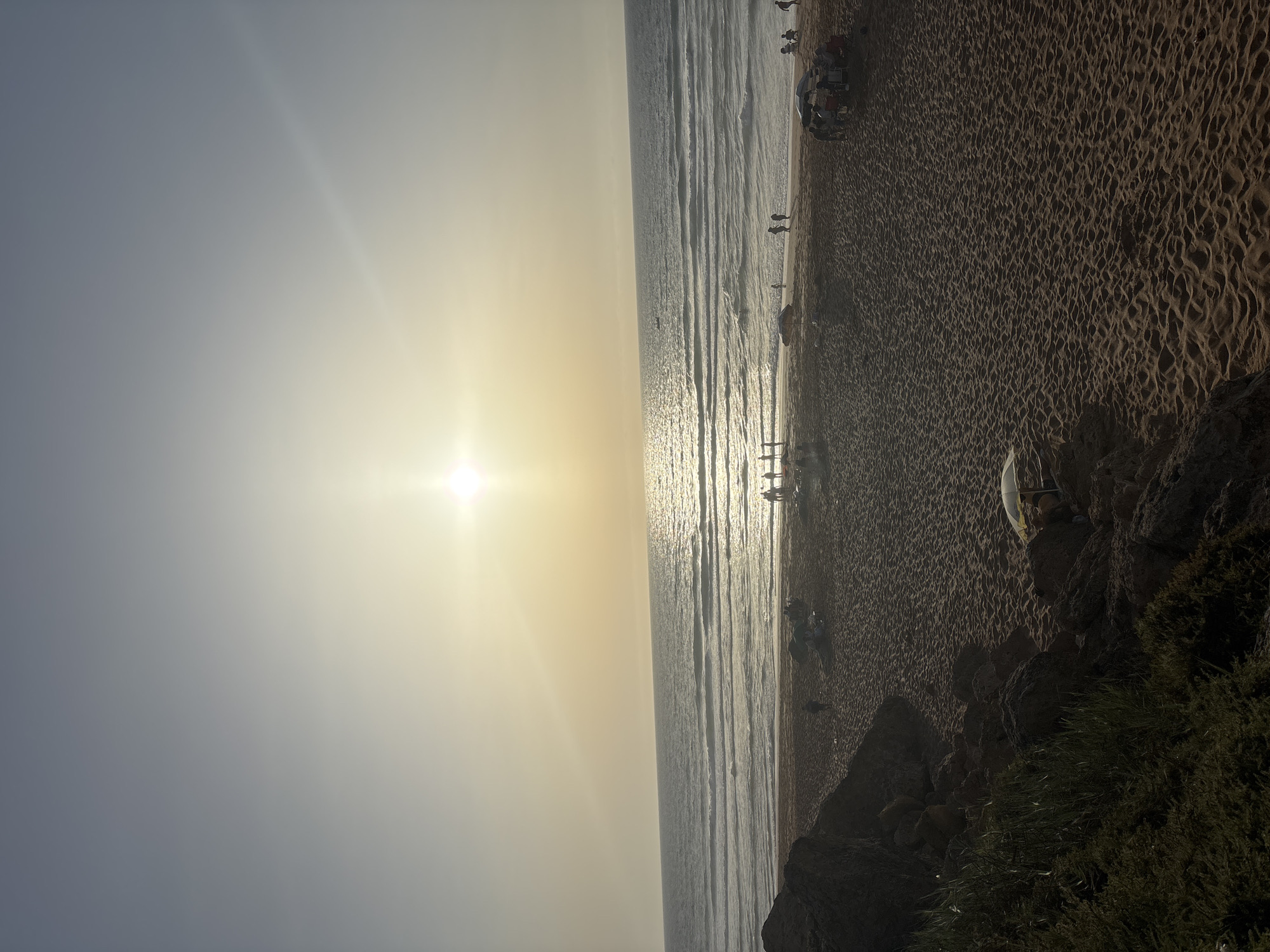 The image depicts a beach scene at sunset. The sun is low on the horizon, casting a warm glow across the sky and reflecting on the ocean. Waves are gently rolling onto a sandy beach where several people are visible, some standing and others gathered around umbrellas. There are also some dark rocks and vegetation in the foreground, suggesting a natural, somewhat rugged coastline. The overall atmosphere is tranquil and serene, typical of a late afternoon or early evening at the seaside.