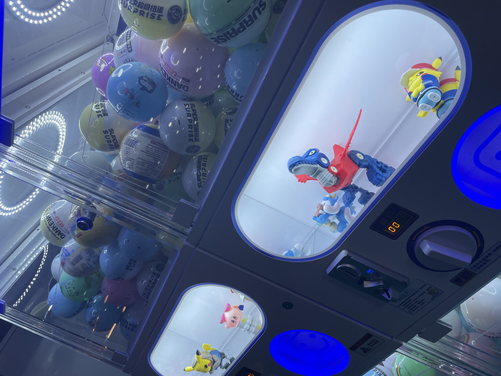 This is a close-up, angled shot of a claw machine filled with colorful, spherical capsules and several small figurines displayed in separate compartments. The machine is brightly lit from within. The upper section is dominated by the capsules, which are predominantly shades of yellow, blue, green, pink, and white. Some capsules have text and graphics, including "SUPER SURPRISE" and what appears to be Japanese writing. Below the capsule-filled area are two illuminated compartments. The left compartment contains two figurines: a yellow character resembling Pikachu wearing a blue hooded jacket and a pink character with a bow. The right compartment contains two toy robots, one predominantly blue and red with visible joints, and another smaller yellow and blue robot. The machine's controls, including a coin slot and a joystick, are visible on the side. The overall environment appears to be an indoor amusement or retail space, likely a mall or arcade. There are no people visible in the frame. The image was taken in Phu Thuy Ward, Vietnam, suggesting this is a public entertainment venue within that location.