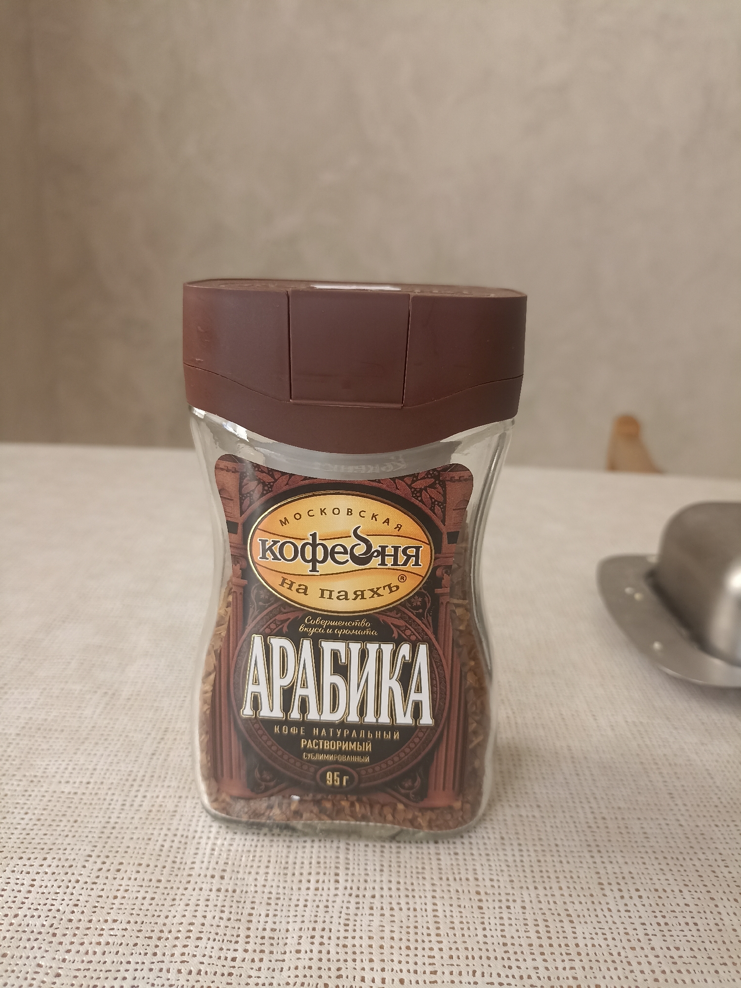 The image shows a glass jar of "Arabika" instant coffee, which appears to be a Russian brand called "Moskovskaya Kofeynya." The jar is brown and transparent, filled with coffee granules. The label on the jar features the brand name prominently, along with text in Russian indicating "coffee on wax," "perfection of taste and aroma," "natural instant coffee," and "freeze-dried." The net weight is stated as "95g." The jar is placed on a white, textured tablecloth. In the background, a plain, light-colored wall is visible. To the right of the jar, a portion of a shiny metal object, possibly a butter dish or lid, is present. There are no people visible in the image. The scene is indoors, with neutral lighting. The location is not specified, and there is no indication it is in the city of Izberbash, Russia.