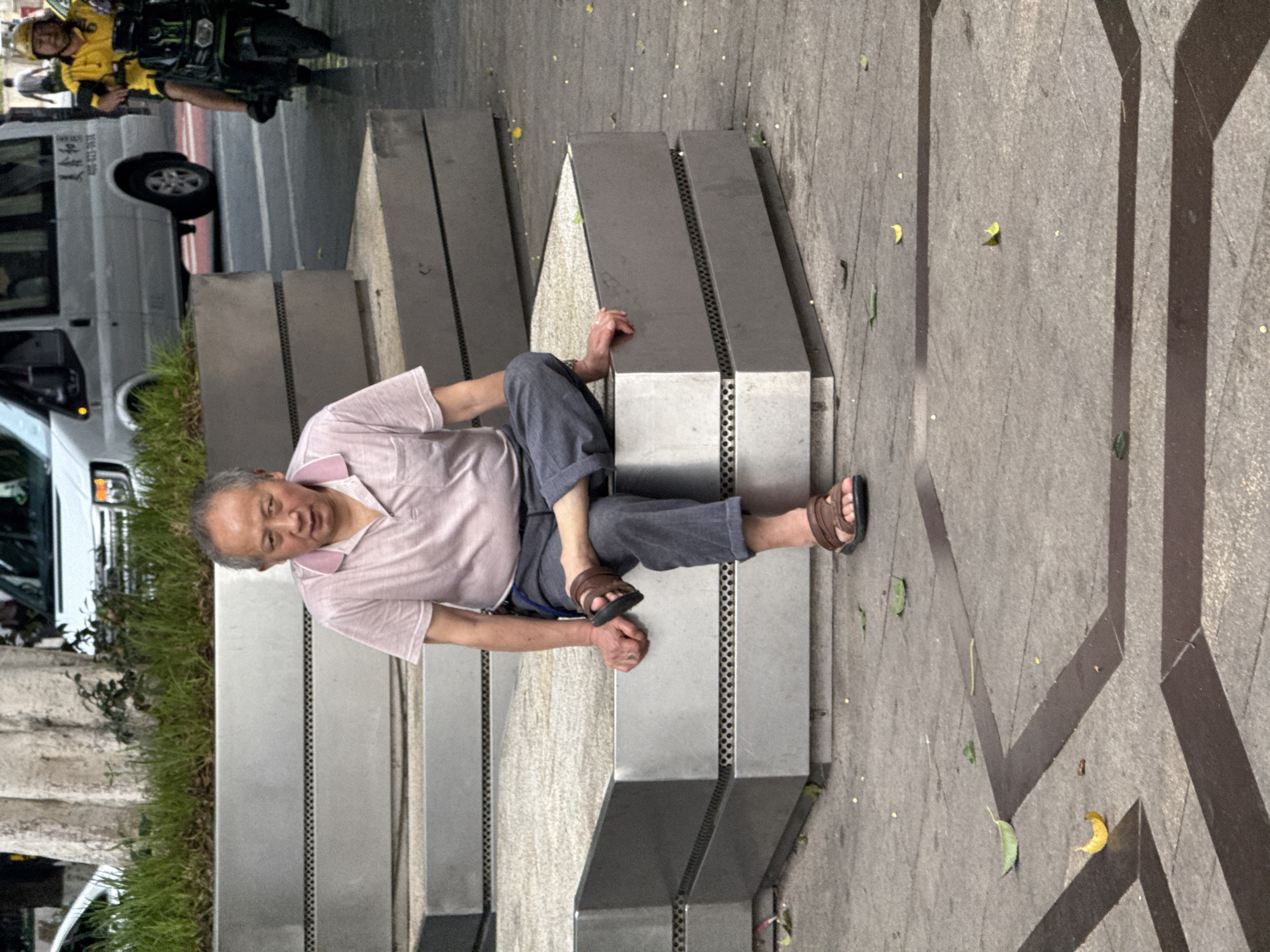 An elderly East Asian man is seated on a modern concrete bench with a metallic facade in Yuzhong District, China. He is wearing a light pink polo shirt and dark gray trousers. His right leg is crossed over his left, and he is wearing brown sandals. His hands are resting on his knees and the bench. In the background, a white van is parked on a wet street, and a person on a motorcycle, wearing a yellow jacket and helmet, is visible. The ground is paved with gray tiles, and scattered leaves are present. The scene appears to be a public urban space.