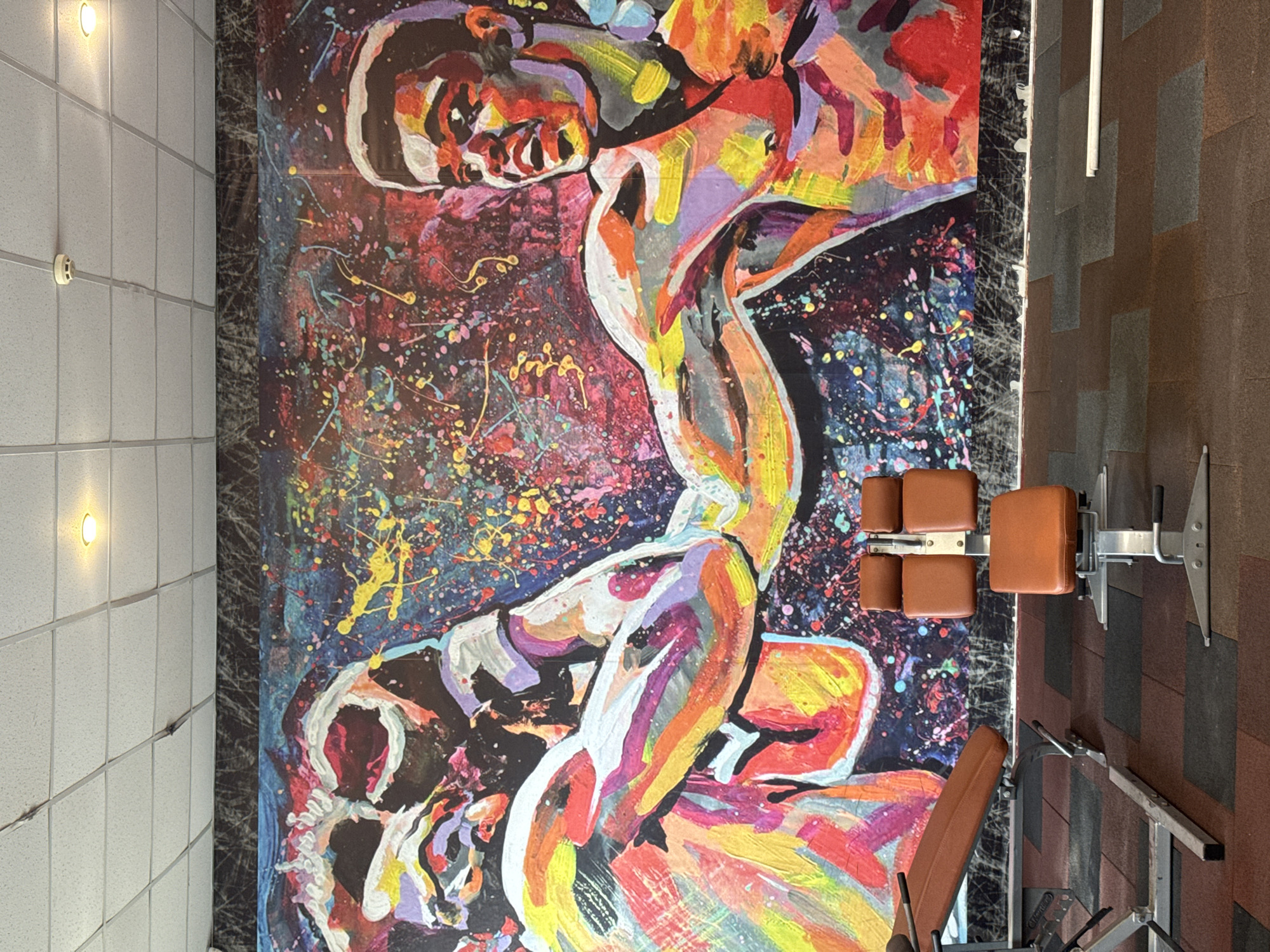 The image shows the interior of a fitness facility in Vladikavkaz, Russia. Dominating the scene is a large, vertically-oriented abstract painting depicting two wrestlers. The painting uses a vibrant color palette of reds, oranges, yellows, purples, and blacks, with splatter effects creating a dynamic background. The wrestlers' bodies are rendered in a highly stylized manner. To the right of the painting, fitness equipment is visible, including weight benches and a leg press machine, suggesting the mural is placed within a workout area. The environment includes a white tiled wall with inset lights to the left and a dark, tiled floor to the right. The overall scene is well-lit.