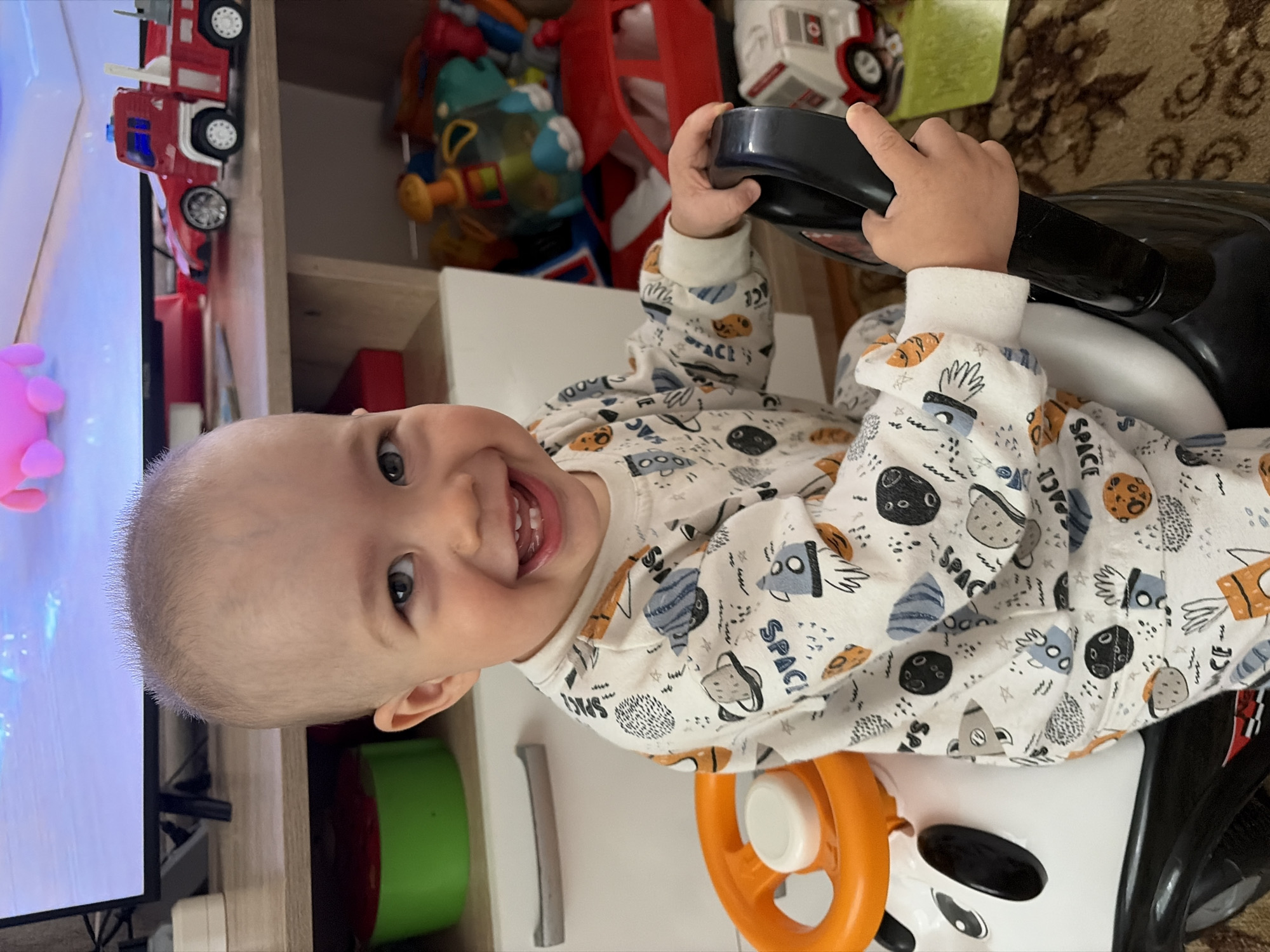 A smiling infant with fair skin and a sparse covering of light-colored hair is centrally positioned in the frame, seated in a child's ride-on toy. The toy features an orange steering wheel and a black exterior. The child is dressed in pajamas with a space-themed print. The background includes a white shelf unit, a television screen displaying a pink hue, and toy fire trucks. Additional toys and items are visible within the shelf unit. The setting appears to be indoors, potentially in a home environment. Although the city is specified, there is nothing in the picture to confirm its location in Chaikovsky, Russia.