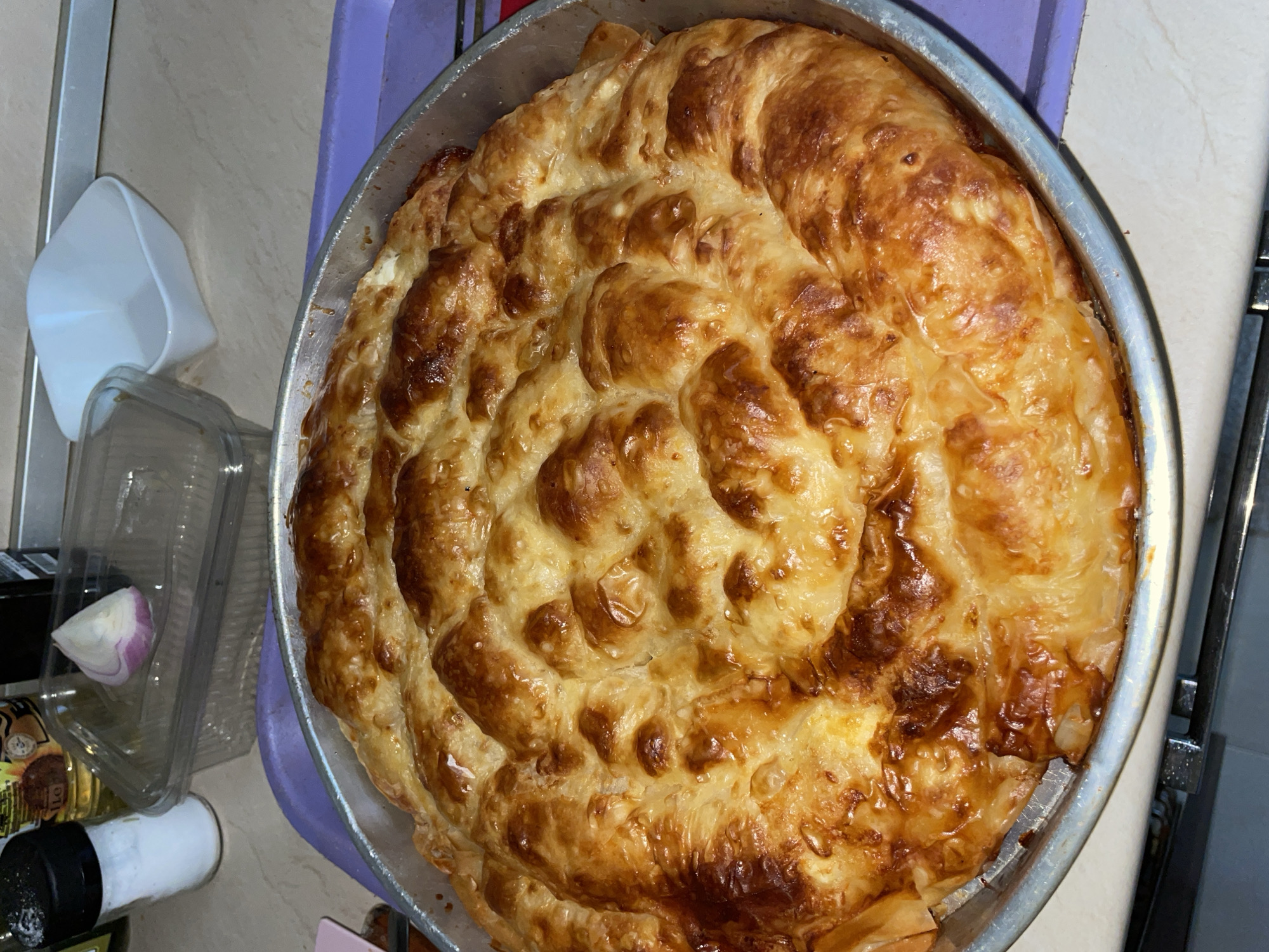 A freshly baked spiral-shaped pastry dominates the frame, nestled within a round silver baking pan. The pastry exhibits a golden-brown hue, indicative of a baked crust. The pan sits on a light purple surface, potentially a tray or countertop. To the left of the pan are various kitchen items, including a partially visible white plastic container, a clear plastic container with a segment of red onion inside, a bottle of oil, and a salt shaker. The scene appears to be indoors, potentially in a kitchen. The location in Zlatograd, Bulgaria is not verifiable from the image.