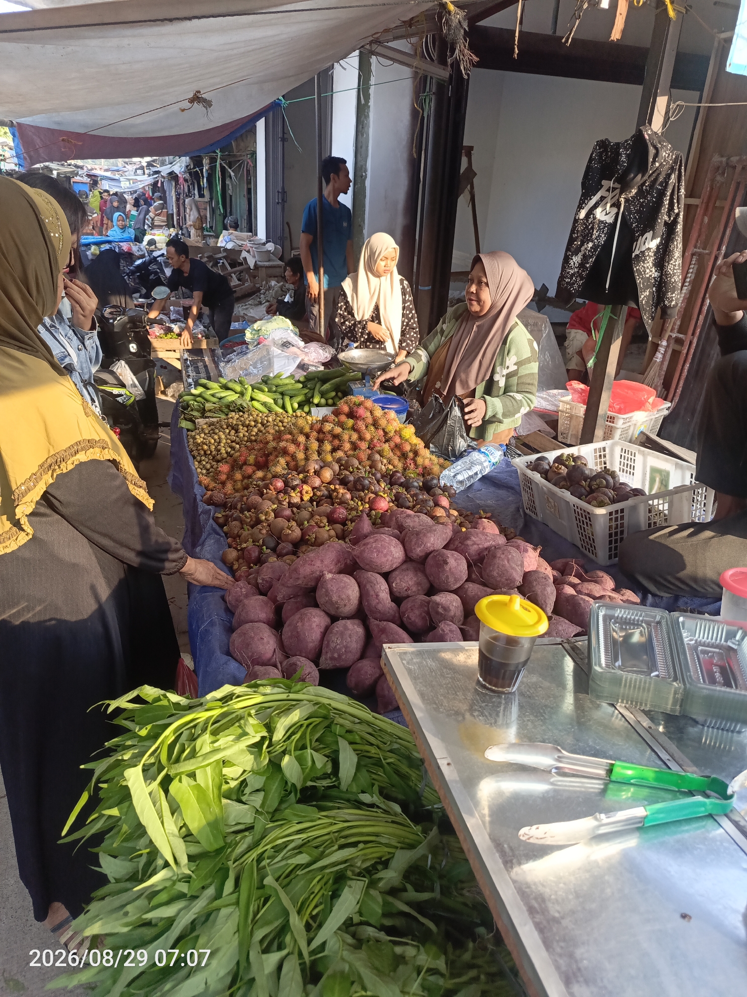 The image depicts a bustling outdoor market scene in Sangiang, Indonesia, likely a local food market. The foreground features a stall with an abundance of fresh produce, including purple sweet potatoes, rambutan, mangosteen, and cucumbers, displayed on blue tarps. Green leafy vegetables are piled high in the lower left corner. A person wearing a yellow hijab and black garment is reaching out towards the produce. To the right, a person wearing a light green sweater and a hijab is engaged in a transaction, holding a plastic bag. Another person in a cream-colored hijab is seated behind a table. In the background, the market extends with various stalls and a walkway, filled with shoppers and vendors. The overall environment appears to be a vibrant and active marketplace.