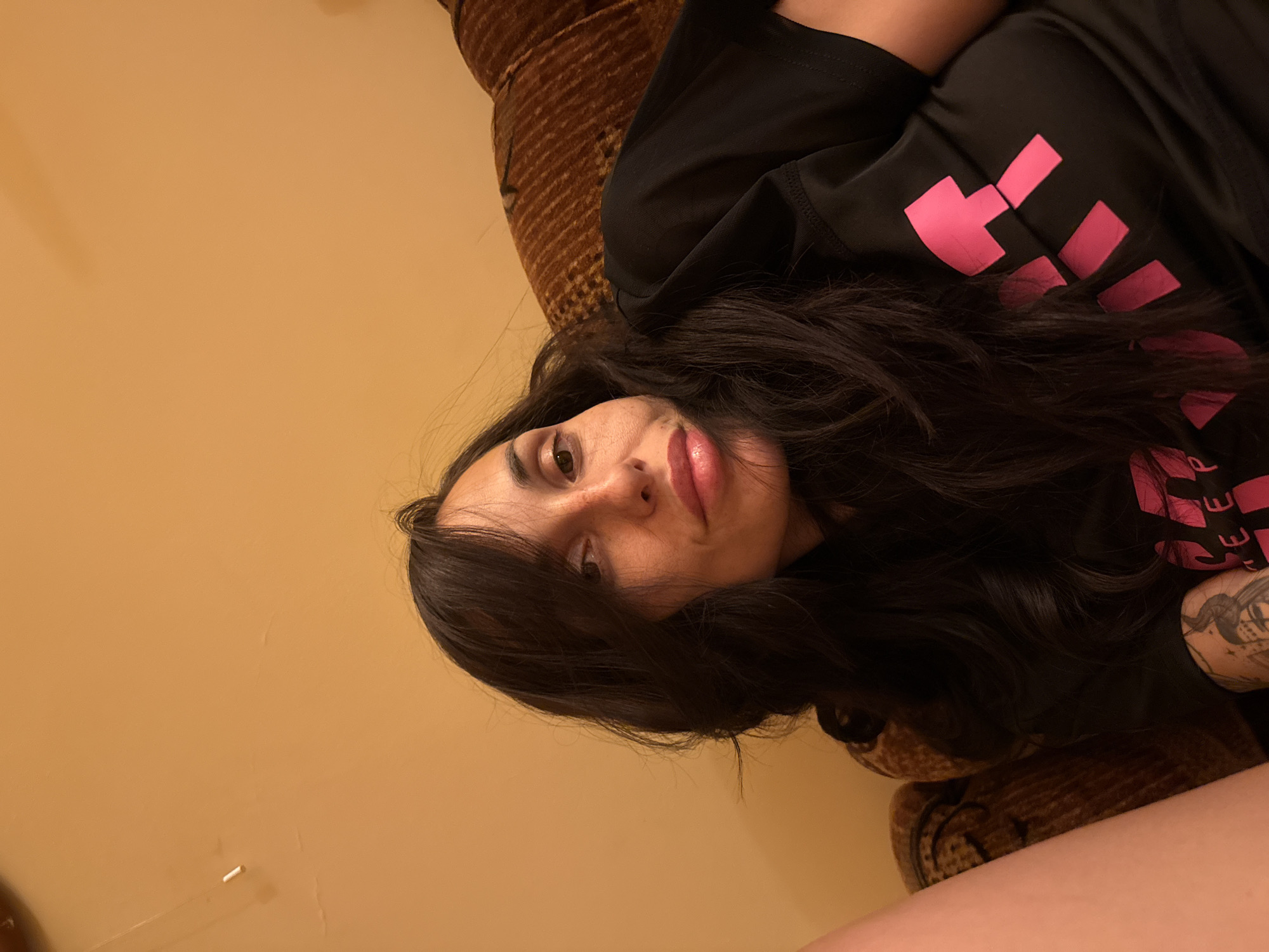 A woman with long, dark hair is lying on a brown upholstered couch, facing the camera. She is wearing a black shirt with pink lettering or symbols. The background is a plain, light-orange wall. The location is potentially within Pleven, Bulgaria, but this cannot be confirmed from the image alone. The image is taken from a low angle. Part of the woman's arm with a tattoo is visible.
