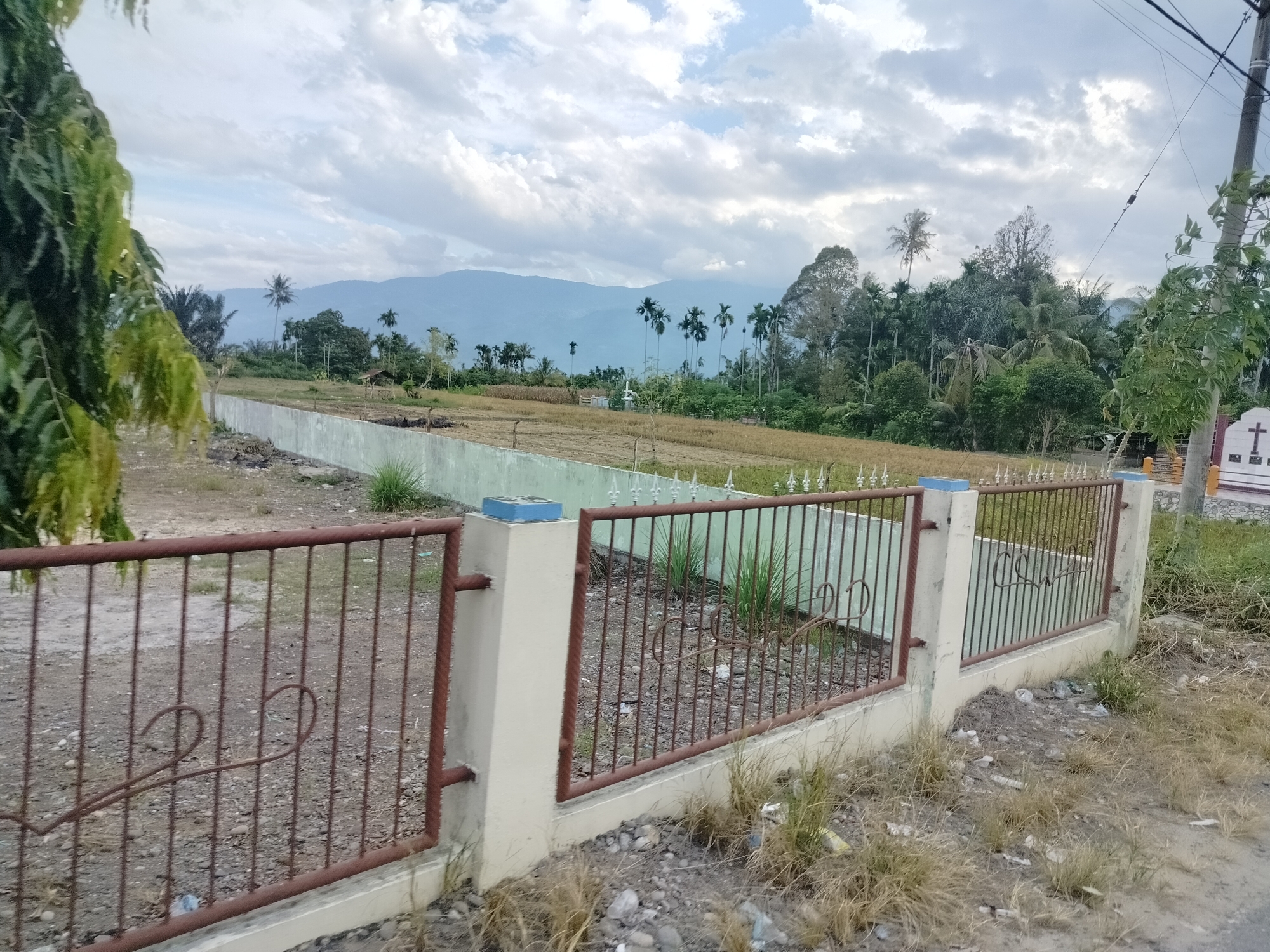 The image depicts a fence in front of a field and a mountain range in the background. The fence is made of metal bars and concrete pillars, with some decorative elements. The field appears to be dry and possibly used for agriculture. In the distance, a lush green landscape with palm trees and mountains under a cloudy sky is visible. The overall scene suggests a rural or semi-rural setting. The context provided indicates the location is Kampungnangka, Indonesia.