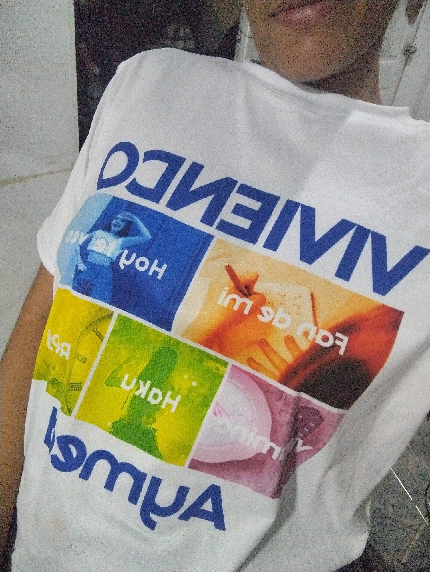 A person is wearing a white T-shirt with a large, colorful graphic print. The print consists of what appears to be a collage of images within rectangular blocks. The words "VIVIENDO" are printed in large blue letters above the graphic. Below the graphic, the letters "MYUA" are also printed in blue. The shirt is partially wrinkled. The person's face is partially visible; specifically the lips, chin, and neck. The background is blurry, but it appears to be an interior space. The location El Valle del Espíritu Santo, Venezuela, is based on the image's metadata, not visual information.