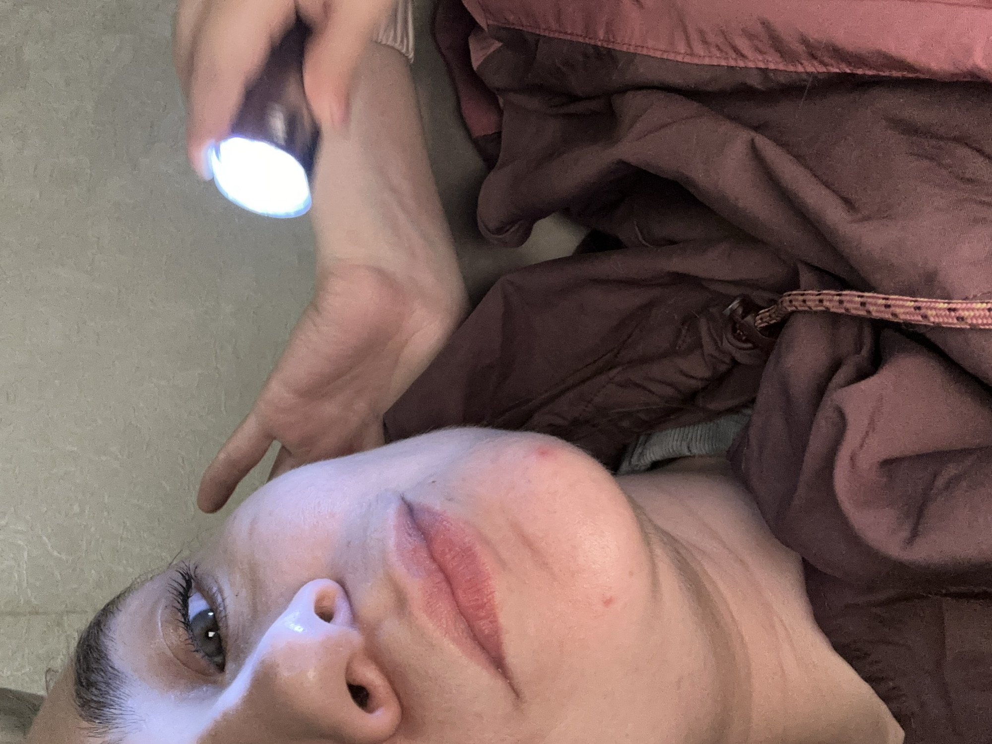 An image from a low angle shows a fair-skinned person lying on what appears to be a light-colored floor. The person has long eyelashes, and their face is angled towards the camera. Their lips are colored, potentially with lipstick, and there is a reddish blemish on their cheek. A hand holding a flashlight is positioned above the person's head, illuminating their face. Part of a dark-colored, possibly maroon, fabric or garment is draped to the side of the person. The image was taken from a tilted angle. No details confirm a location in the city of Uva, Russia.