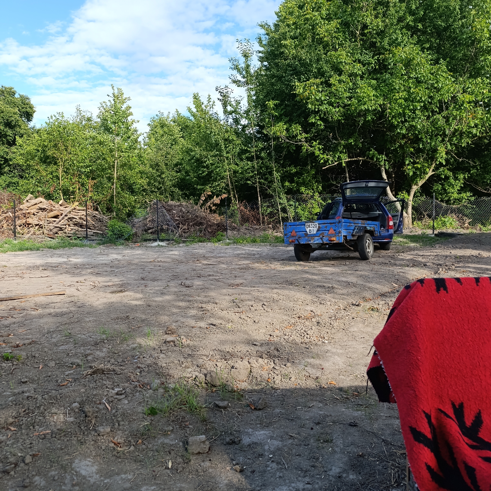 This is a medium-distance, eye-level, outdoor, daylight photograph taken in Ladyzhyn, Ukraine. The scene depicts an open, unpaved area with a blue trailer hitched to a vehicle, partially visible on the right side of the frame. The trailer has a license plate that reads "08320 BI". Behind the trailer, a large tree with green foliage dominates the background, with a chain-link fence running horizontally behind it. To the left of the trailer, a large pile of cut logs and branches is visible, also behind a fence. The ground is predominantly dirt and gravel, with some sparse vegetation and scattered debris. In the foreground, on the right, a portion of a red blanket with a black pattern is visible, obscuring the lower right corner of the image. The sky above is blue with scattered white clouds. No people are present in the frame.
