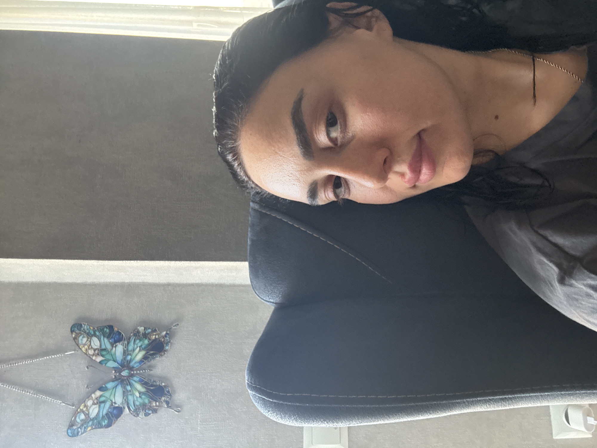 The media file presents an indoor scene, likely in Tbilisi, Georgia, centered on a woman resting her head on a dark, upholstered chair. She appears to be an adult, with dark hair, dark eyebrows, and a calm, possibly relaxed expression as she looks towards the camera. She wears a dark shirt and a delicate gold chain around her neck, with a small mole visible on her skin.
To the left of the woman, on a light-colored, slightly reflective surface, lies a decorative butterfly object. This intricate piece, possibly a pendant or brooch, showcases detailed patterns in various shades of blue and green, and is accompanied by a silver chain.
The background consists of a plain dark gray wall, with a bright light source at the top, suggesting the presence of a window and indicating that it is daytime. The bottom of the image reveals the dark, stitched upholstery of the chair, a light-colored floor, and a white electrical outlet mounted on the wall. There is no visible text in the image.