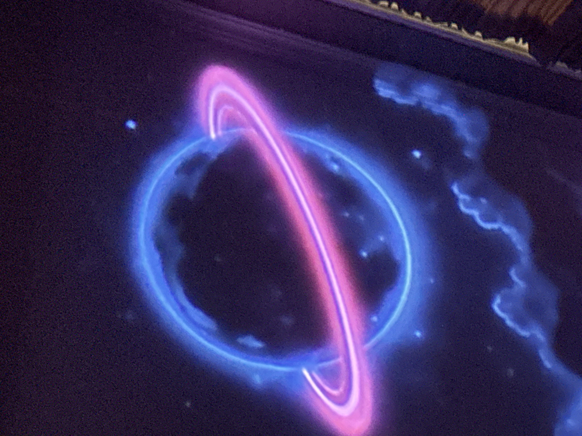 The image depicts a neon sign of a planet with rings, set against a dark background with scattered small lights resembling stars. The planet's glow is rendered in shades of electric blue and vibrant pink, with the rings showcasing a gradient from pink to blue. Subtle cloudy or nebulous forms are visible in the lower part of the image, also illuminated by a faint blue glow. The overall aesthetic suggests a celestial or cosmic theme. The image was taken in Tbilisi, Georgia.