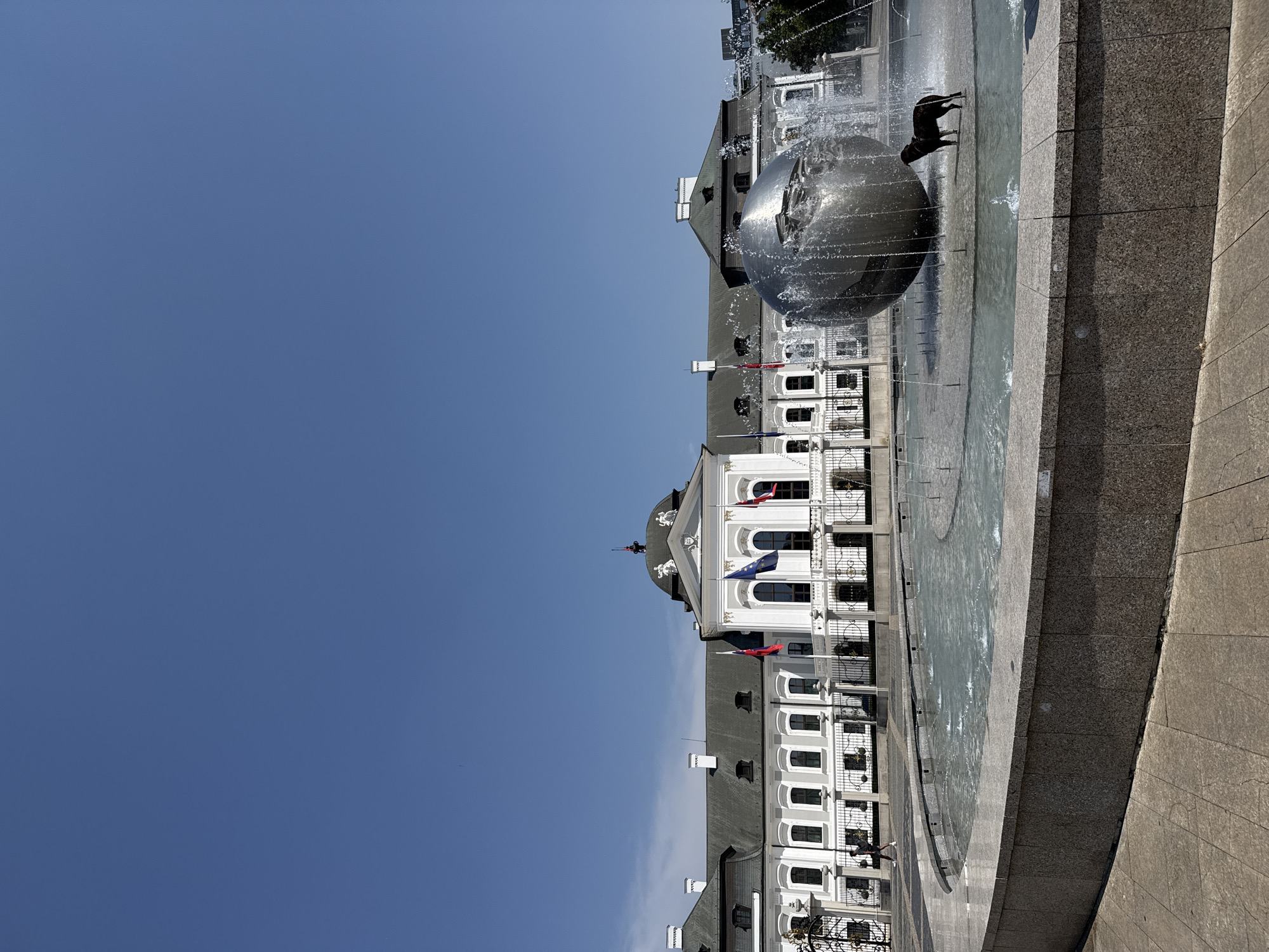 The image shows a wide shot of the Presidential Palace in Bratislava, Slovakia, on a clear, sunny day. In the foreground, a large, modern fountain dominates the scene. The fountain features a large, metallic spherical sculpture at its center, from which water sprays outwards, creating a shimmering effect. A black dog is standing in the shallow water of the fountain, facing to the right.
The Presidential Palace is a grand, white neoclassical building with a central dome. It has a long facade with many windows, a balustrade, and several flags flying from poles in front of it, including the Slovakian flag and the European Union flag. The building is set back from the fountain, with a manicured lawn and fencing in between.
To the left of the palace, a person in dark clothing is walking away from the camera along a paved walkway. The ground in the foreground is paved with stone tiles and concrete. The sky is a clear, bright blue with no clouds visible. The overall impression is one of a public space being enjoyed on a pleasant day.