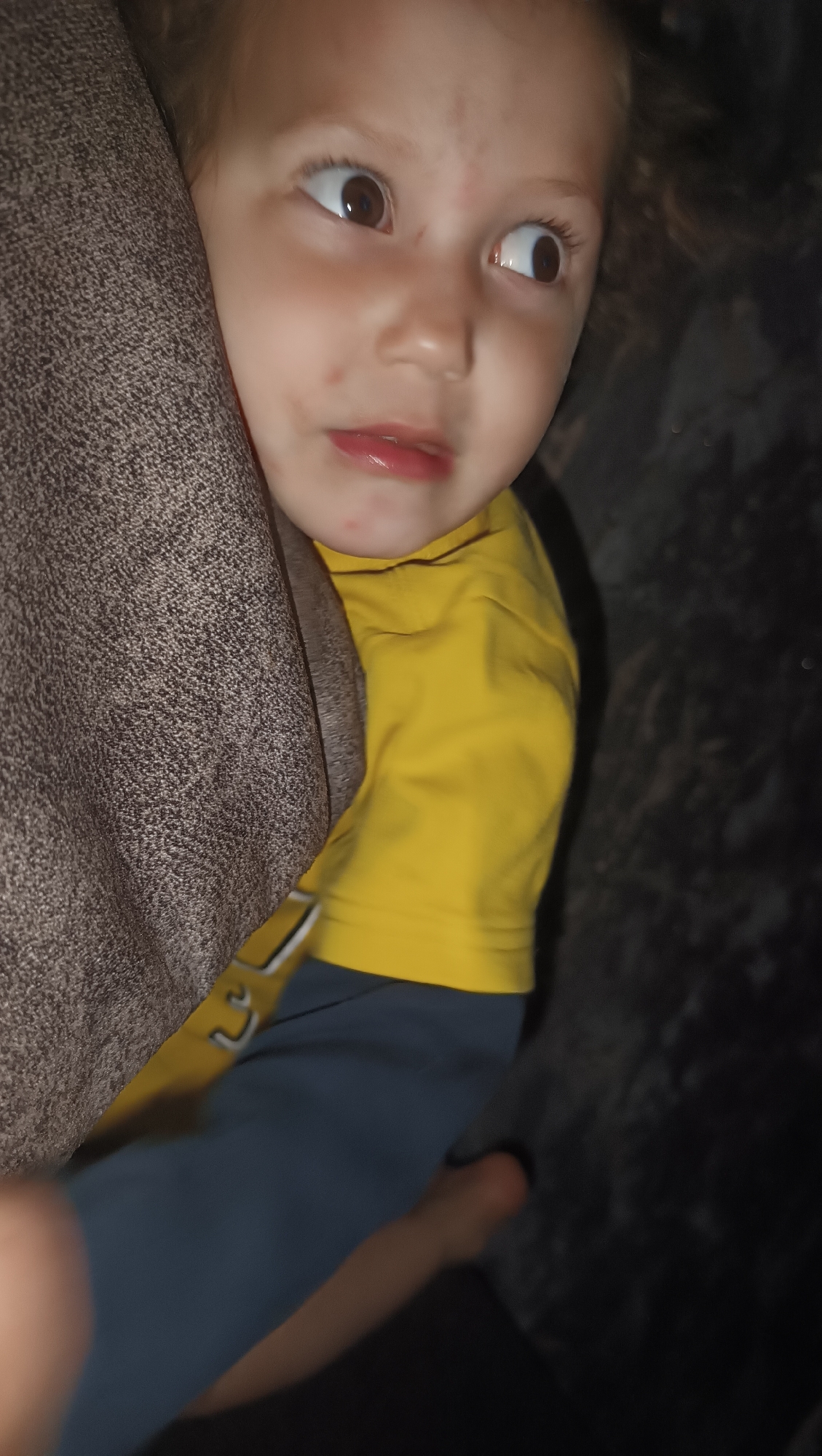 The image shows a close-up of a young child's face, looking to the right with wide, curious eyes. The child has light brown hair and fair skin, with some faint redness on the cheeks and forehead. They are wearing a bright yellow t-shirt. A textured, brownish-grey fabric is partially visible on the left side of the frame, obscuring part of the child's head. The background is dark and appears to be a blurry, textured surface. There is no discernible location in the city of Karsha, Russia, or any other specific environmental context provided by the image.