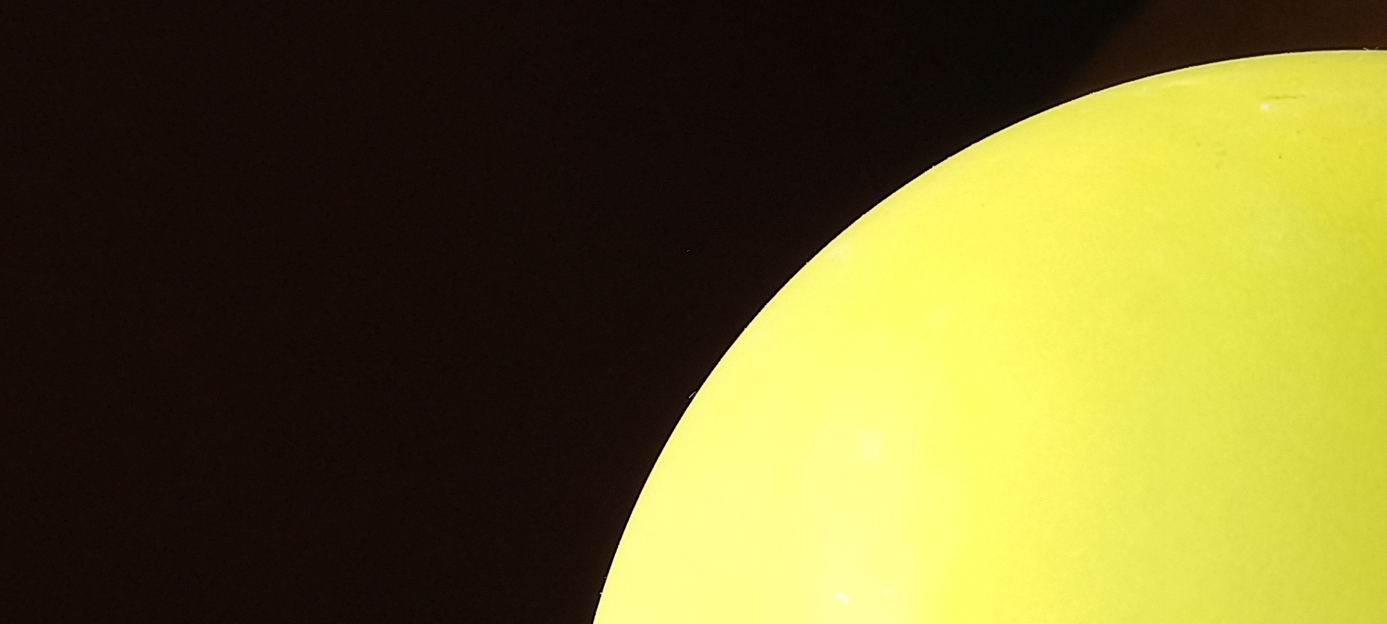 The image displays an abstract close-up, dominated by a large, brightly lit yellow object against a dark, almost black background. The object, occupying the right and lower portions of the frame, appears to be spherical or ovoid, characterized by a smooth, curved edge that sharply contrasts with the void-like background. Its vibrant yellow surface exhibits a subtle matte texture and a few tiny, faint white imperfections. The lighting on the object is soft and even, highlighting its form without revealing specific details about its material or function. The dark background is entirely featureless, providing no environmental context or depth.
There are no identifiable subjects, human or otherwise, nor any specific activity or event taking place. The scene is static, focusing purely on color, form, and the interplay of light and shadow. No visible text is present. Despite the given location context of Kryvyi Rih, Ukraine, the image itself contains no visual cues related to a specific geographical location, time of day, or weather conditions. The composition is minimalist, emphasizing the object's isolated presence and its striking color against the deep darkness.