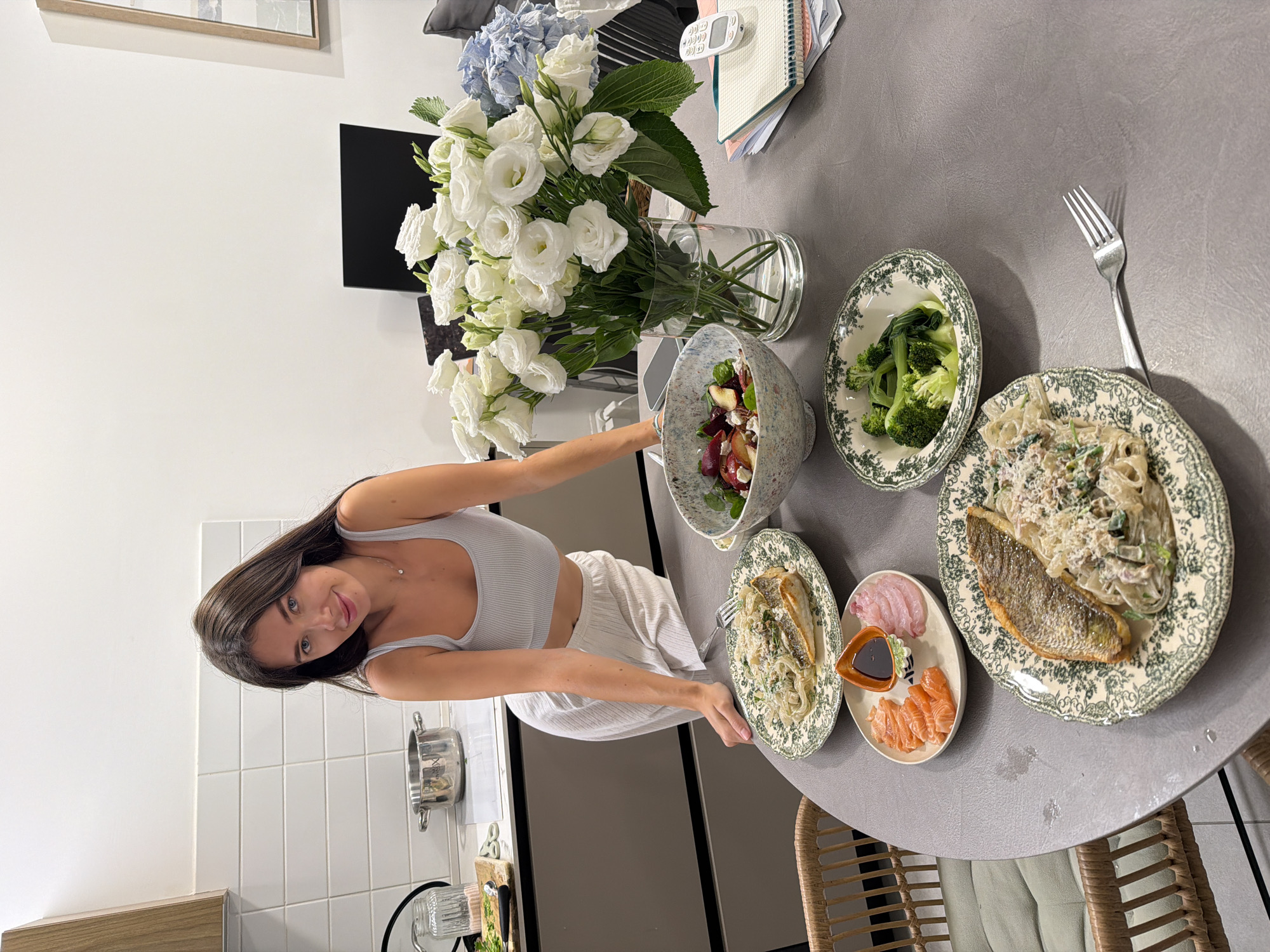 A young woman with long dark hair, wearing a light gray tank top and white wide-leg pants, stands smiling next to a dining table laden with food. She is looking directly at the camera, with one hand near a salad bowl and the other near a plate of pasta and fish. The setting appears to be a bright home kitchen or dining area in Tel-Aviv, Israel.
The gray, textured table is set with a varied meal. A large glass vase in the center holds a prominent bouquet of white Lisianthus flowers and some blue hydrangeas. The food items include two plates of creamy pasta (possibly tagliatelle or fettuccine) topped with a whole pan-fried fish, which resembles sea bream. These plates feature a distinctive green and white floral pattern.
Other dishes visible are a large speckled bowl containing a colorful salad with ingredients such as beetroot, leafy greens, feta cheese, and nuts. A separate smaller bowl, also with the green and white pattern, holds steamed broccoli. Additionally, a rectangular plate offers a serving of sashimi, with slices of salmon and a white fish, alongside a small dish of soy sauce.
In the background, a white tiled wall, a stainless steel pot, and a wooden countertop are visible to the woman's left. Behind the flowers, there's a white wall, a framed picture, a stack of notebooks, and a white remote control. A wicker chair is partially visible in the foreground. The scene is well-lit, suggesting daytime. No visible text is present in the image.