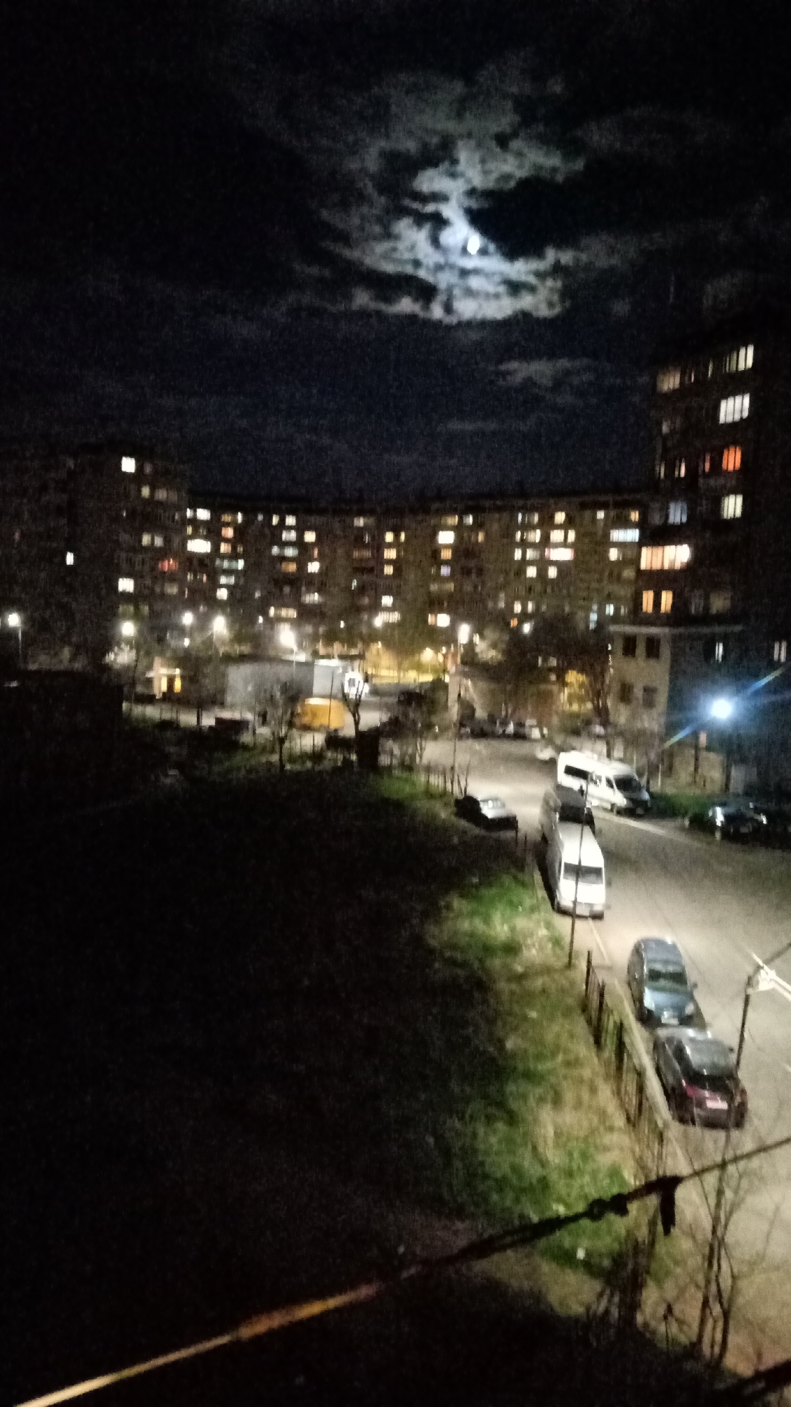 The image depicts a nighttime scene in Yerevan, Armenia, possibly taken from an elevated position overlooking a residential area. In the background, a large, curved apartment building is visible, with many windows illuminated, suggesting occupancy. A second building stands to the right, also lit with windows. Streetlights illuminate the area, revealing parked cars and vans along a road. There is a grassy verge along the road. Above the buildings, the night sky is dark with the moon visible through the clouds. In the foreground, a dark, undefined area is present, possibly a grassy space. A wire is visible in the very bottom of the image.