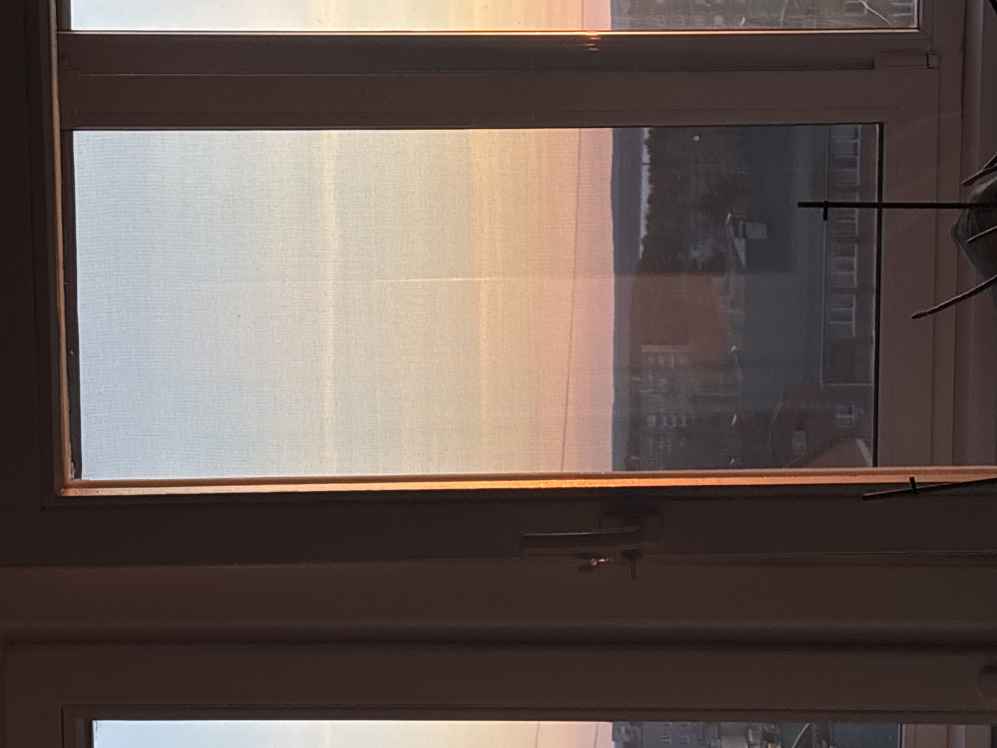 The image depicts a view looking out of a window, which is covered by a translucent, textured screen or sheer curtain. The window frame appears to be white or light-colored, with a dark horizontal mullion separating the upper and lower sections of the window. A window handle or latch mechanism is visible on this mullion.
Beyond the window screen, the sky displays a dramatic gradient of colors, transitioning from light blue at the top to hues of yellow, orange, and soft pink towards the bottom, indicative of either sunrise or sunset. A dark, indistinct horizon line separates the colorful sky from a faint, distant landscape or cityscape below, where muted outlines of multi-story buildings are discernible, particularly on the right side.
An external, thin, dark horizontal rod or antenna is visible extending from the right side, outside the building, across the view. Inside the room, to the far right, part of a green, leafy houseplant is visible, suggesting an indoor setting. There is no specific activity taking place, only a serene view. No text is visible in the image. The scene conveys a tranquil atmosphere, captured at dawn or dusk in Chaikovsky, Russia.