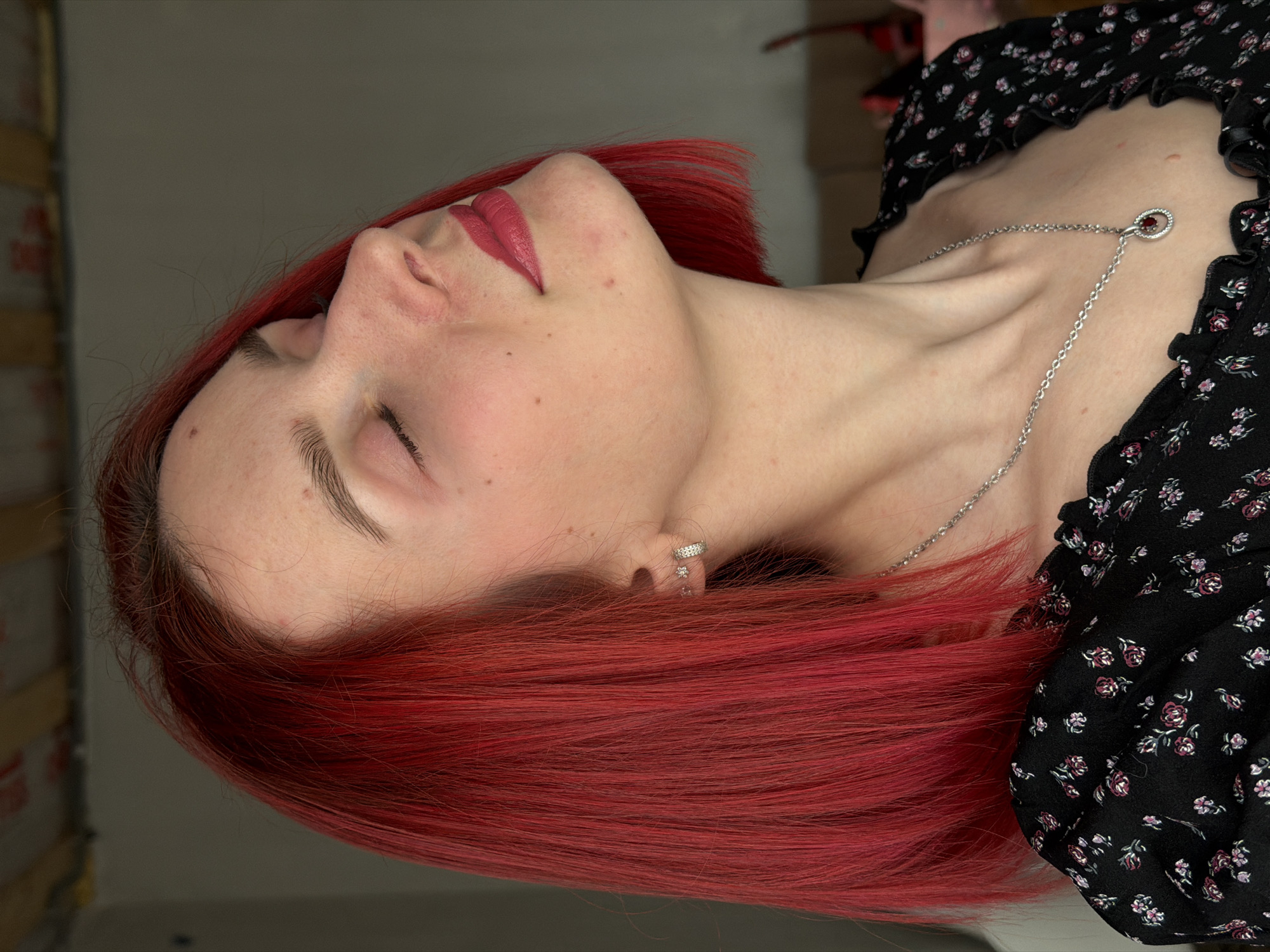 A person with red hair is shown in a close-up, tilted view. The person's eyes are closed and face is turned upwards. The person is wearing a black top with a floral pattern and a silver necklace with a small pendant. The background is indistinct, suggesting an indoor location. The location is in the city of Chaikovsky, Russia.