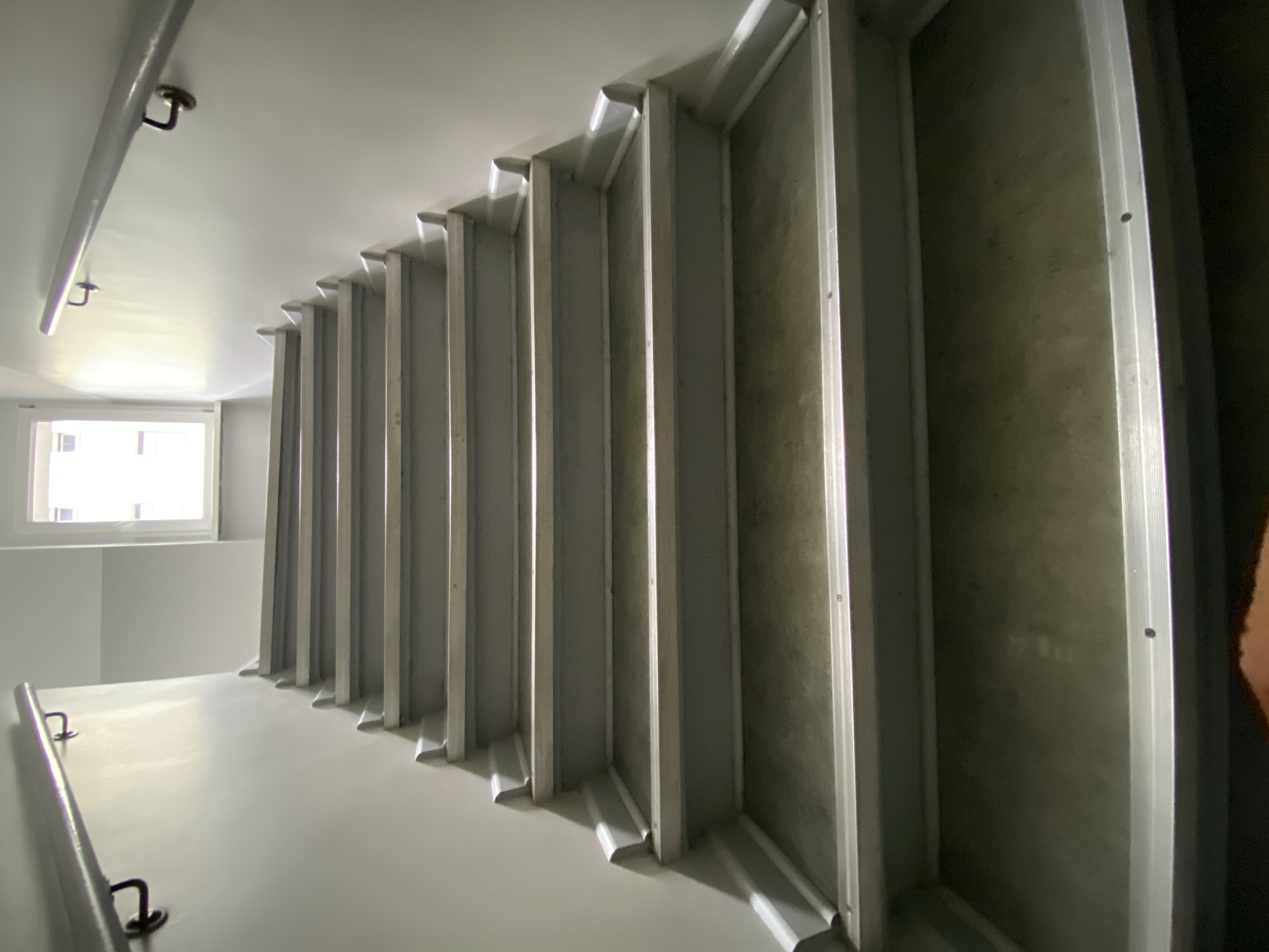 This media file shows a view looking up a metal staircase in what appears to be an apartment building or a similar interior space. The subjects are the staircase itself, constructed from corrugated metal or a similar material for the steps, and the white walls of the stairwell. There are also metal handrails visible on the left side of the frame, attached to the wall with brackets. In the background, a window with white panes lets in natural light, suggesting it is daytime. The overall scene is minimalist and functional, with a focus on the architectural elements of the stairwell. The lighting is bright and airy near the window, becoming more shadowed further up the stairs. The location cues are minimal, but the mention of Szczecin, Poland, provides geographical context. No people are visible in the image, and no specific activity is taking place. There is no visible text. The mood is neutral, conveying a sense of utilitarian architecture.