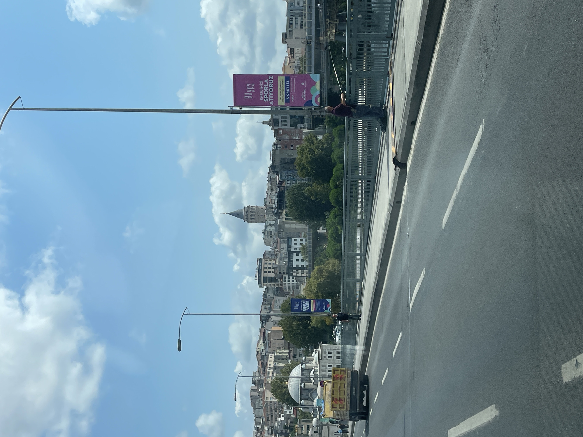 The image shows a view of the Galata Tower in Beyoğlu, Turkey, from a bridge. In the foreground, a two-lane road with white lane markings is visible. A yellow truck is traveling in the left lane. On the right side of the road, a pedestrian walkway runs alongside a metal fence. A person is fishing from the walkway, holding a fishing rod. A large pink banner with Turkish text is hanging from a lamppost on the walkway. In the middle ground, there is a modern building with a dome, possibly a mosque, to the left, and a complex of city buildings with a variety of architectural styles. Trees are visible between the buildings. The Galata Tower is a prominent landmark in the background, seen above the city skyline. The sky is blue with scattered white clouds.