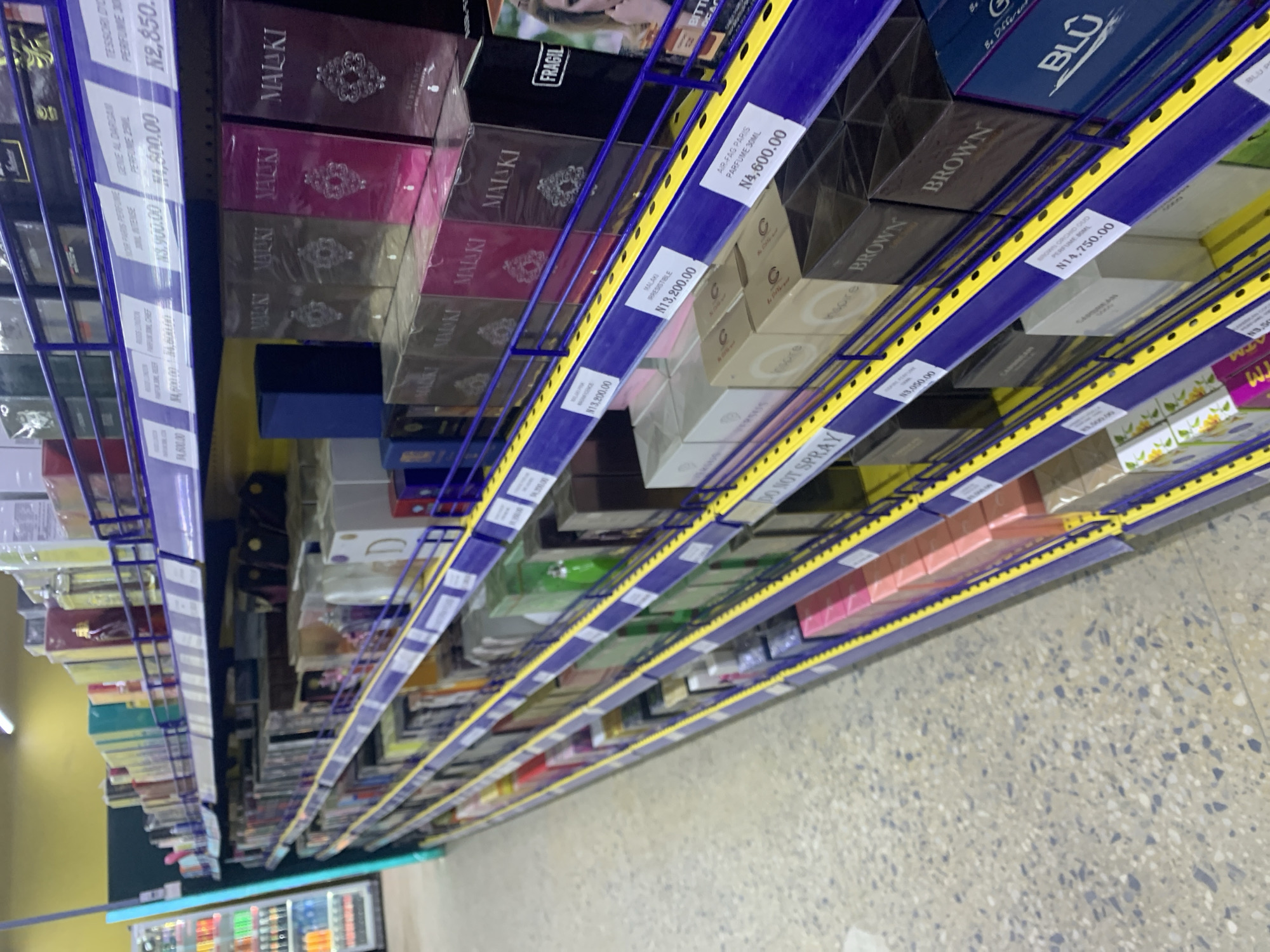 The image depicts a retail environment, specifically an aisle in a store selling perfumes. The shelves are stocked with numerous perfume boxes, predominantly in darker shades of packaging like black, brown, gold, and pink, with some lighter colored boxes visible as well. Several shelves are labeled with prices and product names, such as "MALAKI," "BROWN," and "BLU," along with numerical values in Naira (N). The shelves are made of metal and have a purple and yellow color scheme. The flooring is a terrazzo-style pattern. In the background, a glimpse of refrigerated beverage displays can be seen to the left, suggesting this is a supermarket or a department store with a dedicated fragrance section. The scene is illuminated by overhead lighting.