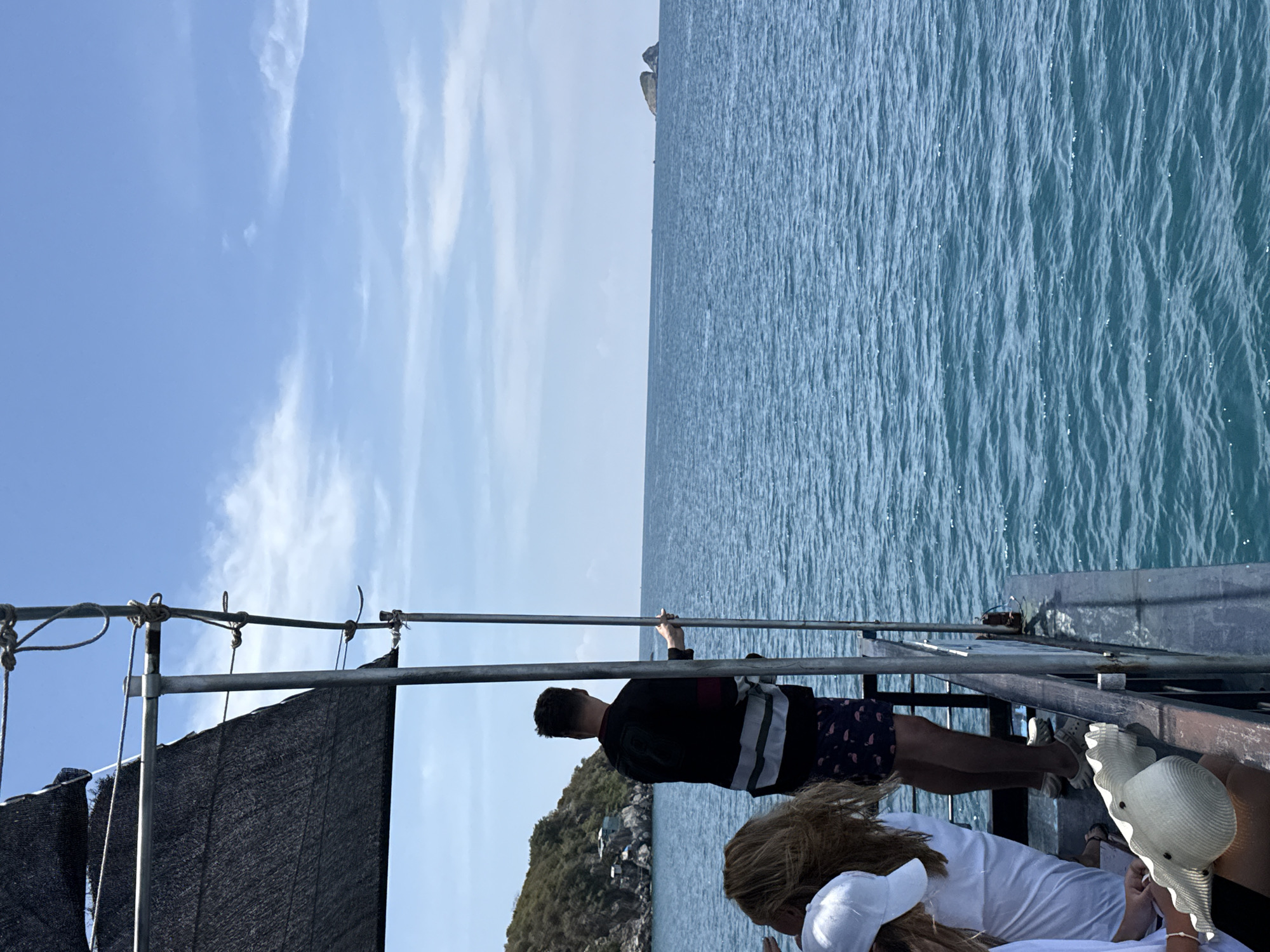 On a boat in what appears to be the city of Bac Nha Trang Ward, Vietnam, a man in a black and white striped shirt and patterned shorts stands at the railing, looking out over the blue water. Another person in a white shirt and hat sits nearby. The sky is blue with scattered clouds.
