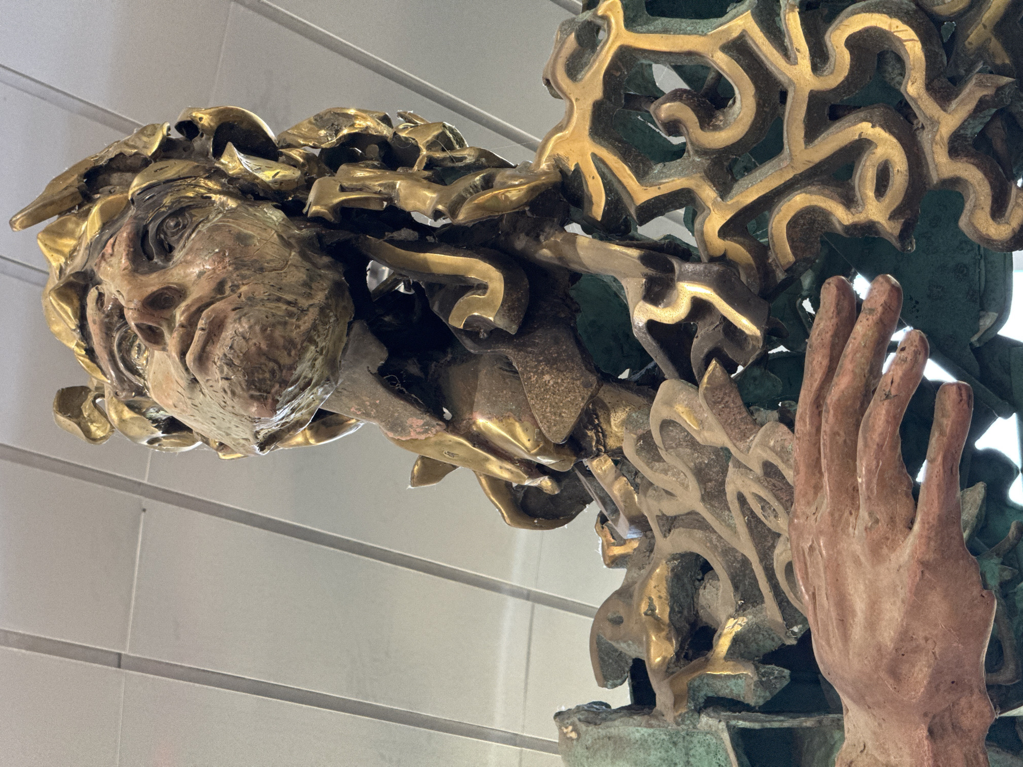 This close-up image showcases a highly detailed and ornate multi-material sculpture. The central subject is a stylized human face, likely male, depicted in a golden-toned metal such as bronze or brass. The face features expressive contours, a prominent eye, nose, and a textured mustache or beard, with intricate, flowing elements resembling hair or decorative flourishes surrounding it.
To the right of the face, an open, sculpted hand appears in a reddish-brown material, possibly copper or patinated bronze. Both the face and hand are intertwined with an elaborate structure of swirling, organic, and geometric metallic patterns. These patterns exhibit a striking contrast between highly reflective golden sections and areas covered in a greenish patina, indicative of aged bronze. The sculpture's intricate craftsmanship highlights varying textures and a rich material palette.
The background consists of a light-colored, possibly paneled or tiled, wall, suggesting an indoor setting like a museum or a public space. The sculpture is well-lit, emphasizing its metallic sheen and the depth of its intricate details. No activity is taking place as it is a static art installation. No text is visible on the sculpture or in the background. This artistic piece is located in Sofia, Bulgaria.