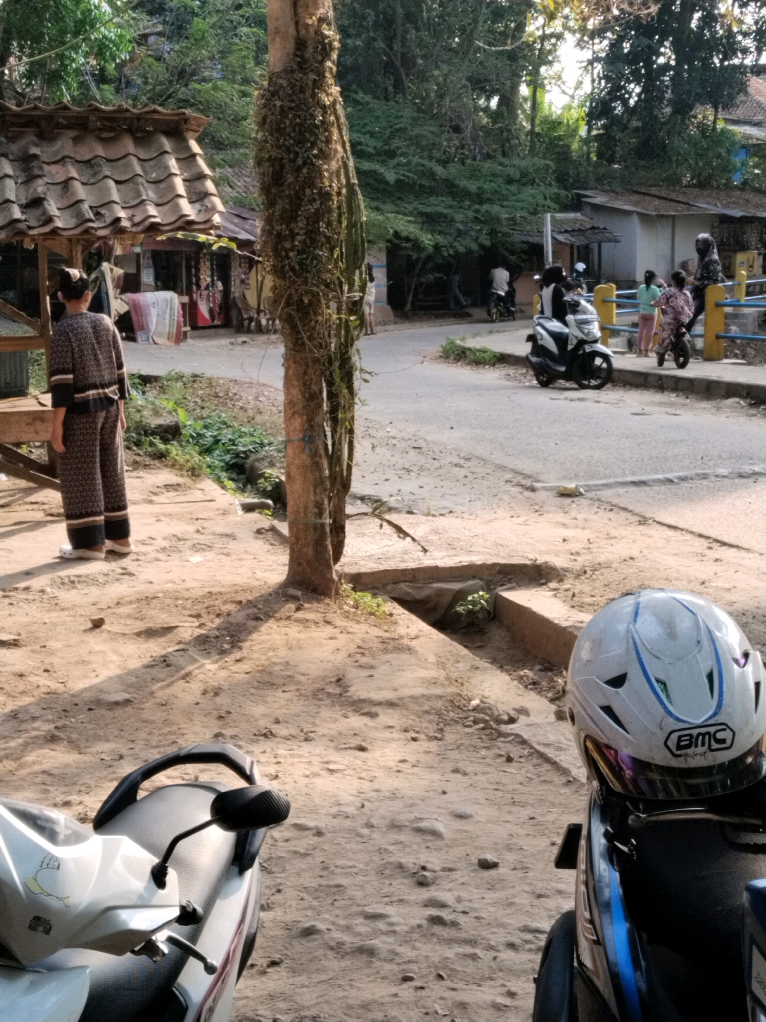 A medium-shot, eye-level, vertical photograph depicts a sunlit, dusty outdoor scene in Ciomas, Indonesia. In the foreground, a white motorcycle is parked on a dirt path, with its seat and handlebars visible. A white helmet with blue accents and a "BMC Helmet" logo rests on the motorcycle's seat. A large, rough-barked tree with vine growth stands to the left of the motorcycle. In the middle ground, a person wearing a patterned dark outfit stands with their back to the camera, facing a small open-air structure with a tiled roof. This structure appears to be a shop, with items displayed inside. To the right, a paved road is visible, with a yellow and blue metal railing running alongside it. Several individuals, including children, are present near the railing, some standing and one on a small motorbike. Another motorcycle is parked on the road in the background. The environment is characterized by dirt paths, a paved road, trees, and simple buildings, suggesting a semi-urban or rural setting in Ciomas. The lighting suggests it is daytime, possibly late afternoon, with strong shadows cast by the tree.