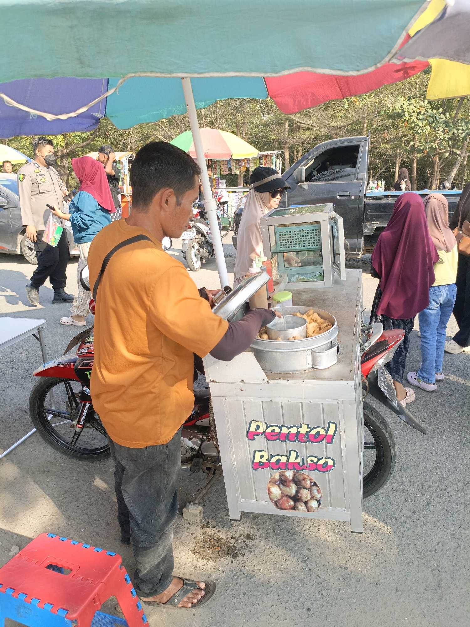 In Bulukumba, Indonesia, a street vendor sells "pentol bakso" from a cart, identifiable by the signage on its side. The scene, likely a market or roadside stall, features a variety of individuals. A man in an orange t-shirt and dark pants, presumably the vendor, is attending to the food cart, which holds a large metal pot filled with fried food items. He is seen using tongs to serve customers.
Several people are present, including a woman in a light-colored hijab and a black hat, wearing sunglasses, who appears to be a customer or observer. Another woman in a dark red hijab and jeans stands nearby, as does a person in a yellow shirt and blue jeans. A uniformed police officer in a mask is also visible, observing the surroundings. The background reveals a typical urban environment with trees, a parked vehicle, and other stalls or shops. Colorful umbrellas provide shade over the vendor's cart. The ground is paved with asphalt, and a red and blue plastic stool is placed to the left of the cart. The overall impression is of a busy daytime scene at a local food vendor.