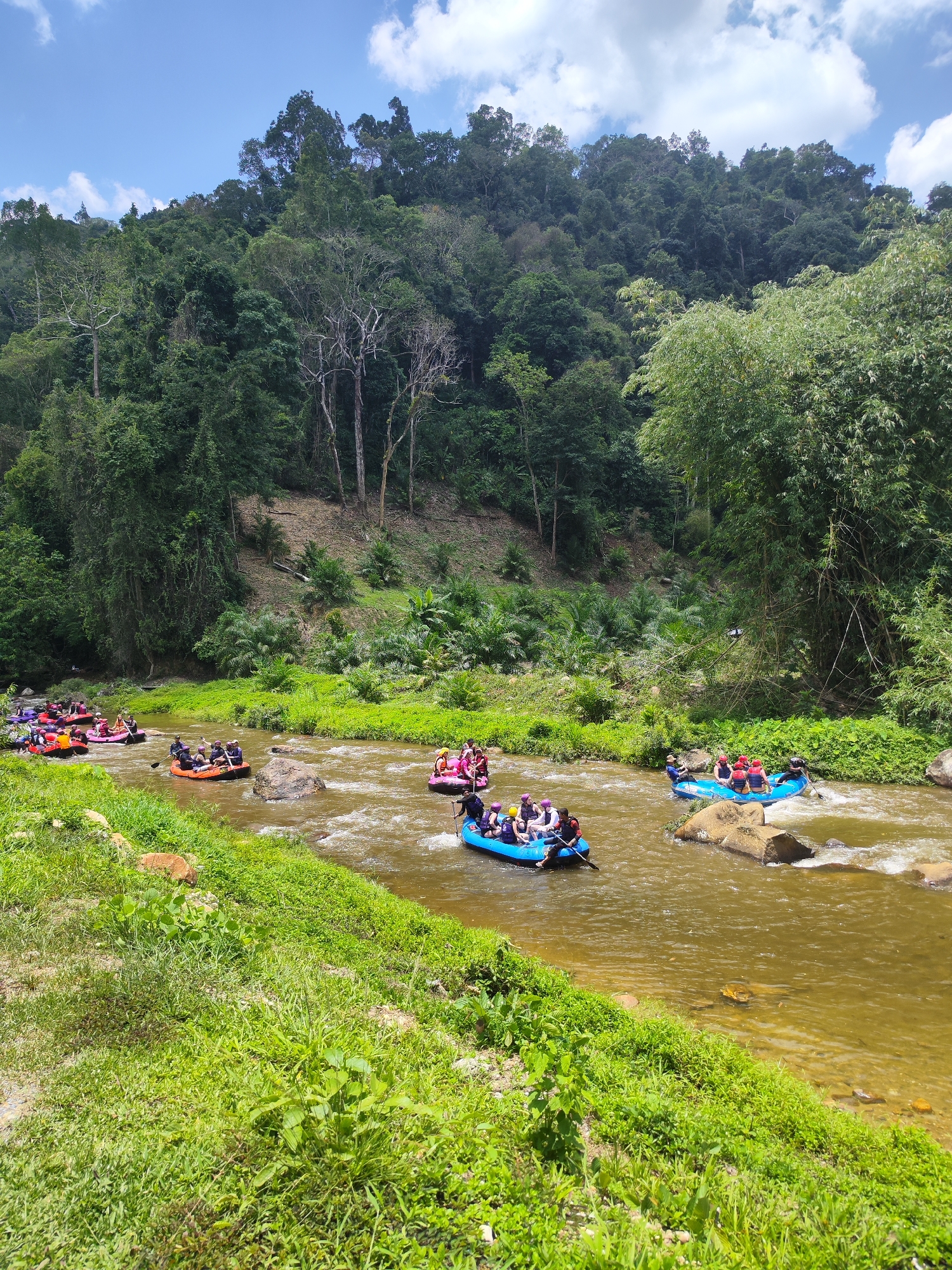 The image depicts a group of people participating in a white water rafting activity on a river in Tambon Song Phraek, Thailand. Several inflatable rafts, each carrying multiple individuals, are navigating the river's current. The river water is a murky brown, and rocks are visible both within and along the riverbanks. Lush greenery, including various trees and vegetation, covers the surrounding landscape and rises up the slopes in the background. The sky is blue with some white clouds.