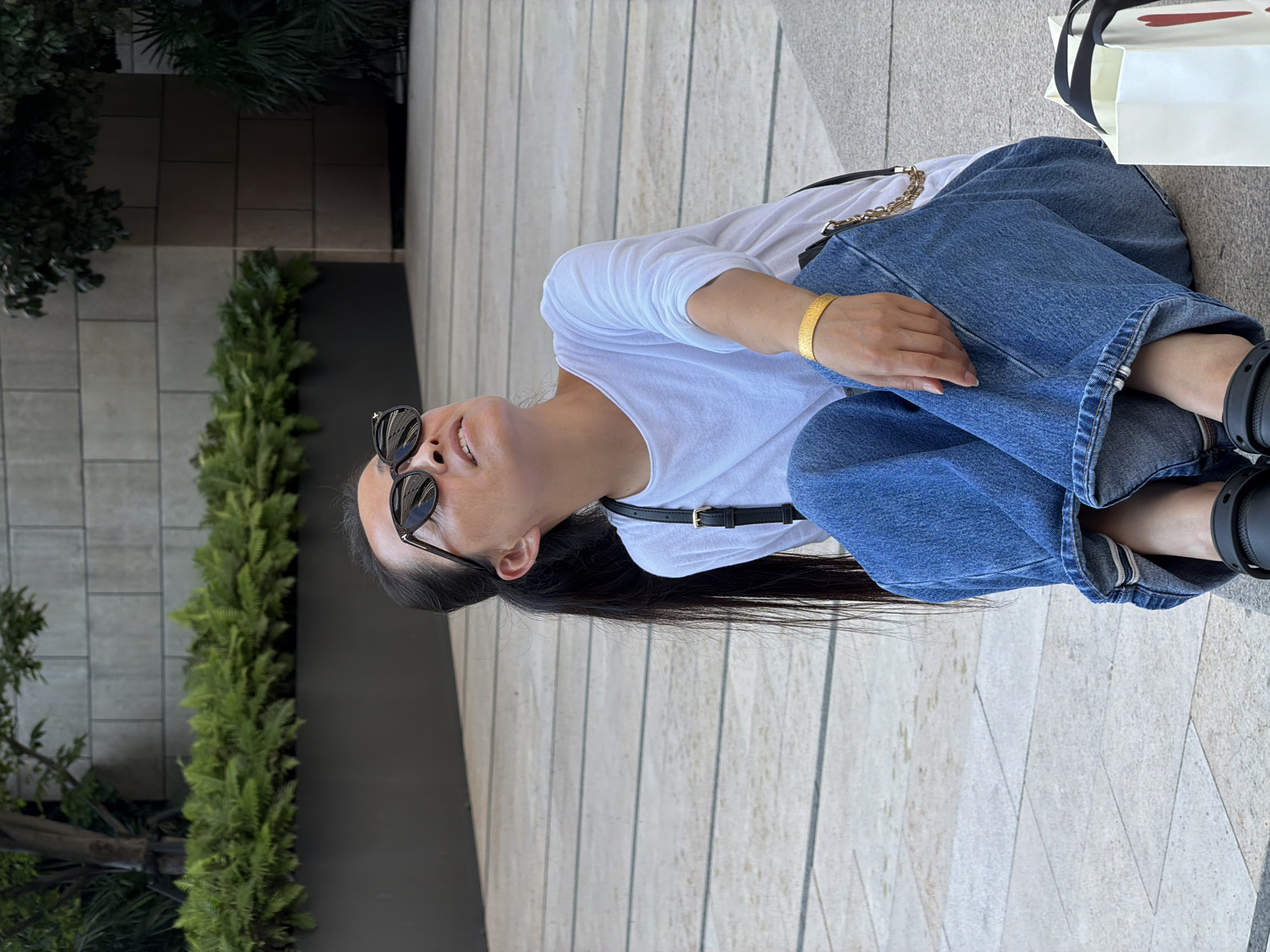 An Asian woman wearing blue jeans, a white t-shirt, and sunglasses sits on a stone ledge outdoors. She is looking upwards and to her right. A black strap with a gold chain is visible across her shoulder. A white shopping bag with a red heart design is next to her. The background consists of a paved area and a planter with green plants and trees, with a building facade behind it. The location is identified as Yuzhong District, China.