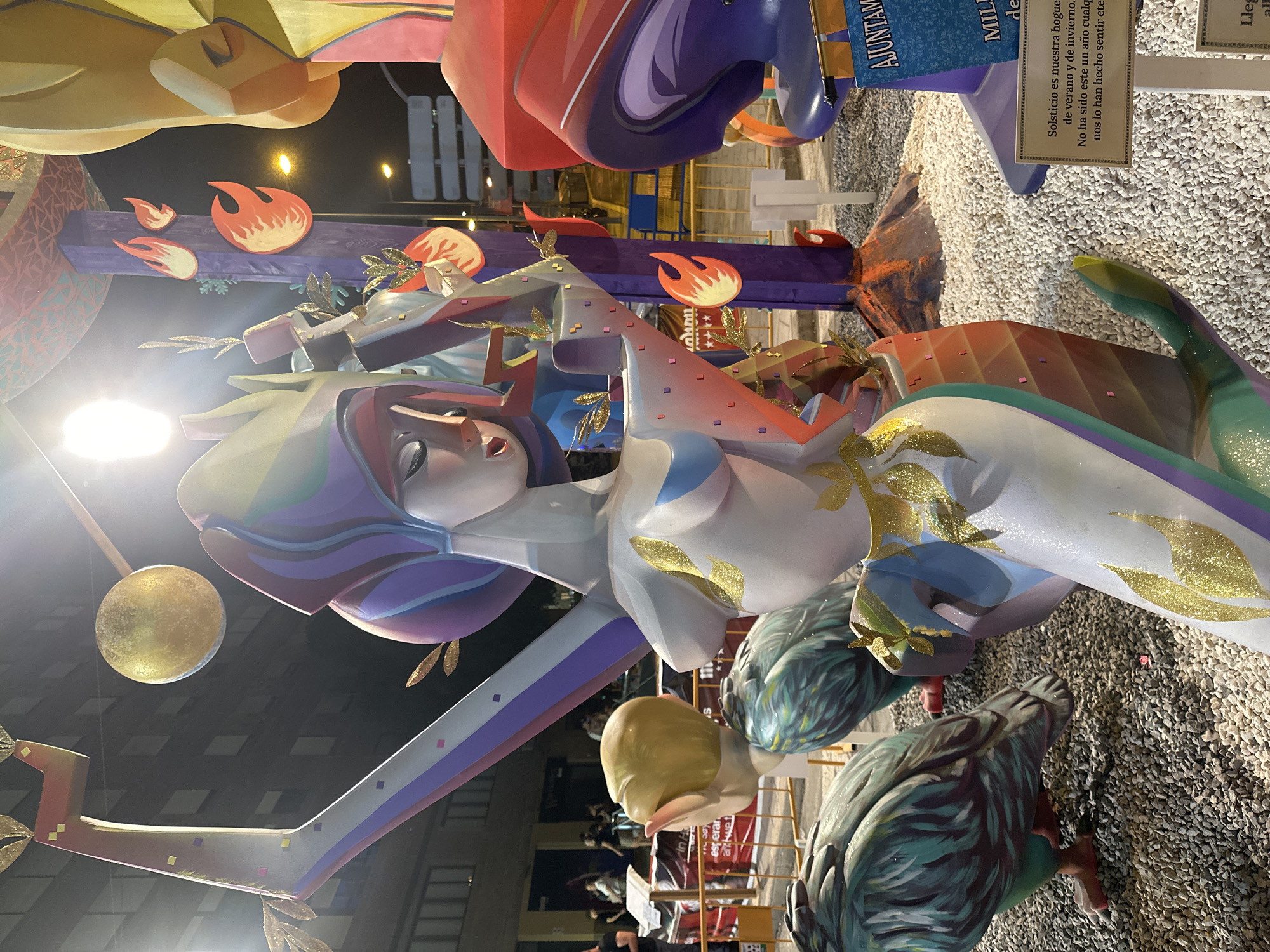 The image shows a nighttime outdoor scene featuring elaborate, colorful sculptures that appear to be part of a festival or parade. The sculptures are made of a material resembling papier-mâché or painted wood and depict stylized figures and decorative elements. In the foreground, a large sculpture of a woman with a flowing headdress and a white, glittery garment adorned with gold leaf details is prominent. To the left and slightly behind her are two large, feathered bird-like sculptures. The background includes other large, abstract sculptures in bright reds, purples, and yellows, as well as illuminated streetlights and buildings. A street sign with the text "AJUNTAMIENTO DE ALICANTE" is visible in the lower right corner, indicating the location is Alicante, Spain. The ground is covered in white gravel. Several people are visible in the background, suggesting the presence of an event with attendees. The overall impression is festive and artistic, likely related to a local celebration in Alicante.
