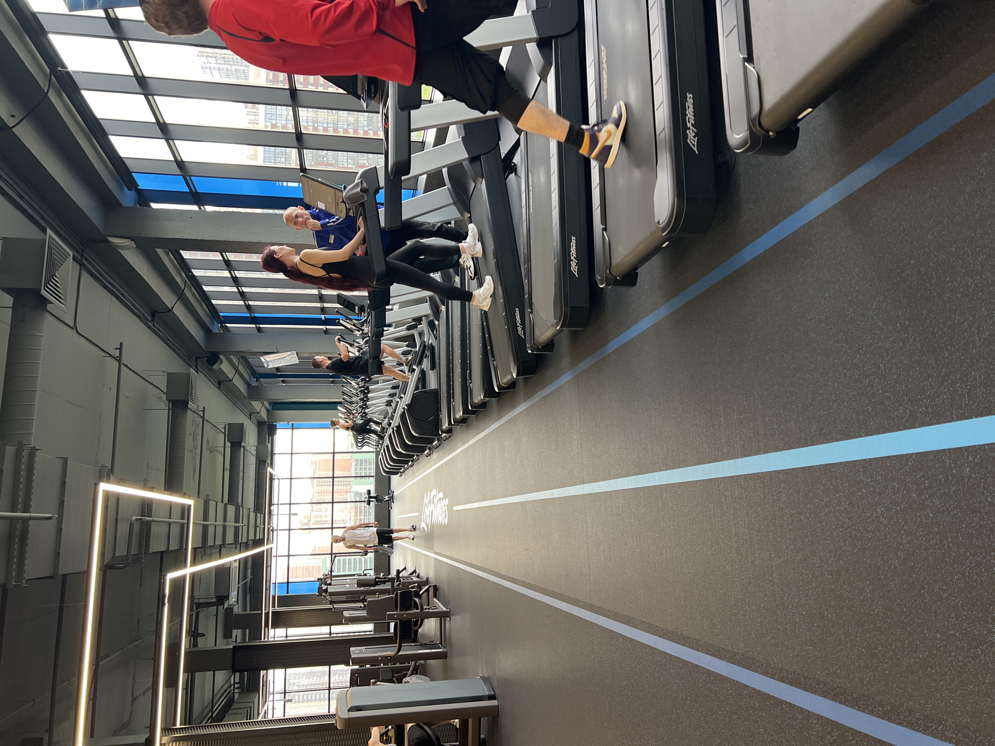 The image captures a fitness center setting, featuring a row of Life Fitness treadmills occupied by individuals engaged in exercise. A running track is marked by blue stripes running down the center of the room. The architecture of the building includes a high ceiling with a grid of windows providing natural light and views of the city, specifically buildings that suggest a Krasnodar, Russia, location. The room is equipped with lighting fixtures and various exercise machines along the walls. People are walking or running on the treadmills and some are walking near the weight machines, indicating a mix of cardiovascular and strength training activities.