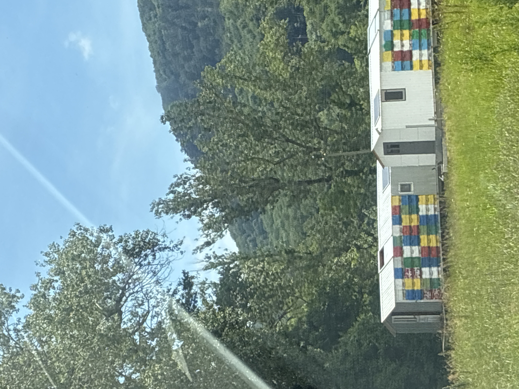 An exterior, eye-level, medium shot shows a colorful, long, modular building, possibly a beekeeping structure, in the city of Tekos, Russia. The building has a metal roof and is covered with what appear to be bee hives, painted in red, blue, yellow, and green squares. Two small windows and a dark-colored door are visible on the structure. The building sits on a wooden platform in a grassy field. Behind the building, a lush green forest covers a hillside under a bright blue sky with faint white clouds. Trees in the foreground obscure parts of the sky and the far left side of the building.
