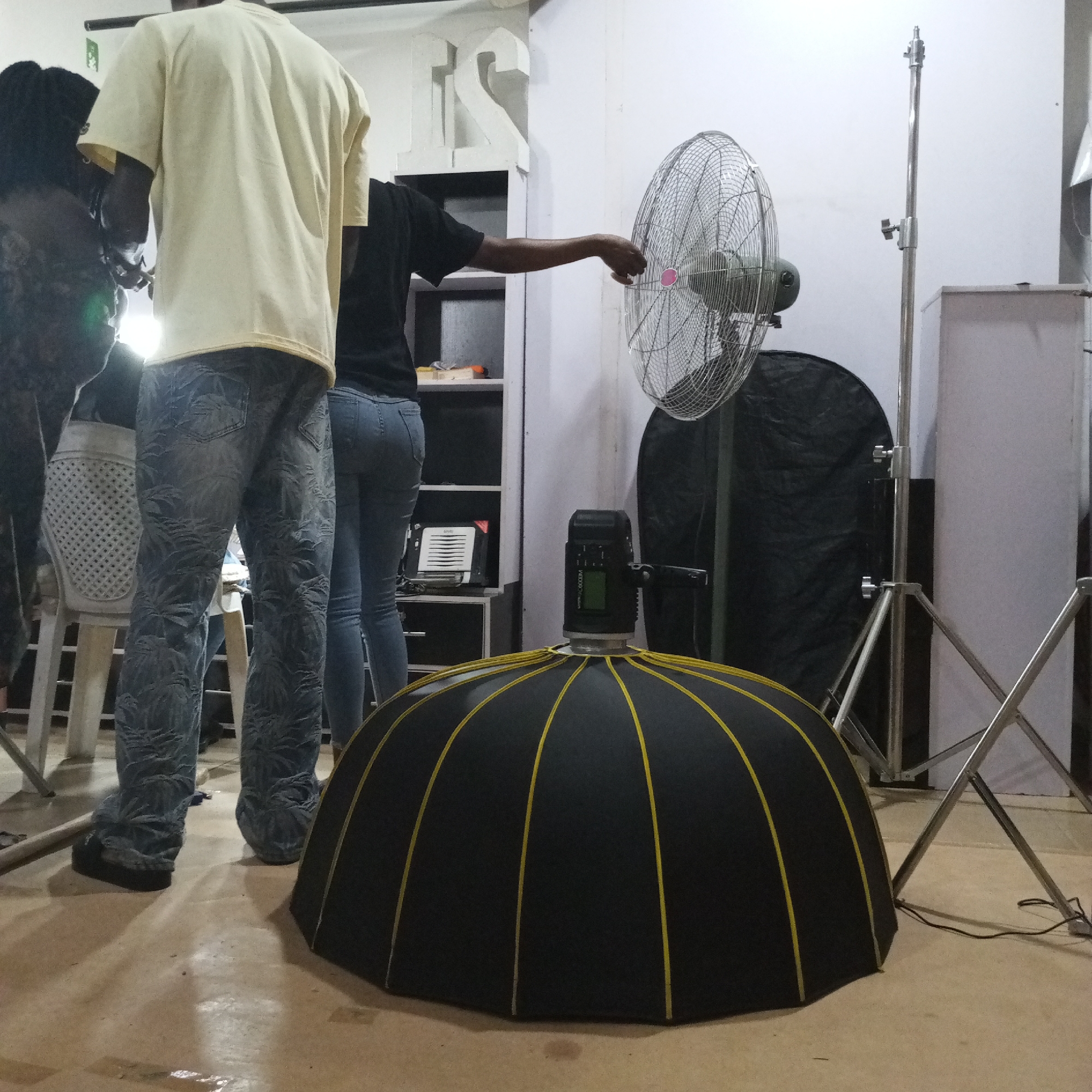 A photograph taken in what appears to be a studio setting, potentially in Bukuru, Nigeria, features a mix of people and equipment. The foreground is dominated by a large black dome-shaped softbox with yellow ribs. A studio light is attached to the top of the softbox. Behind it, a standing fan is placed, with a person in a black shirt seemingly adjusting it. To the left, two other individuals are present, one wearing a yellow shirt and patterned jeans, and another partially visible person. The background includes a light stand, a black photography reflector, and a storage unit with shelves. The floor is a light brown or beige color. The scene suggests a photography or videography shoot in progress.