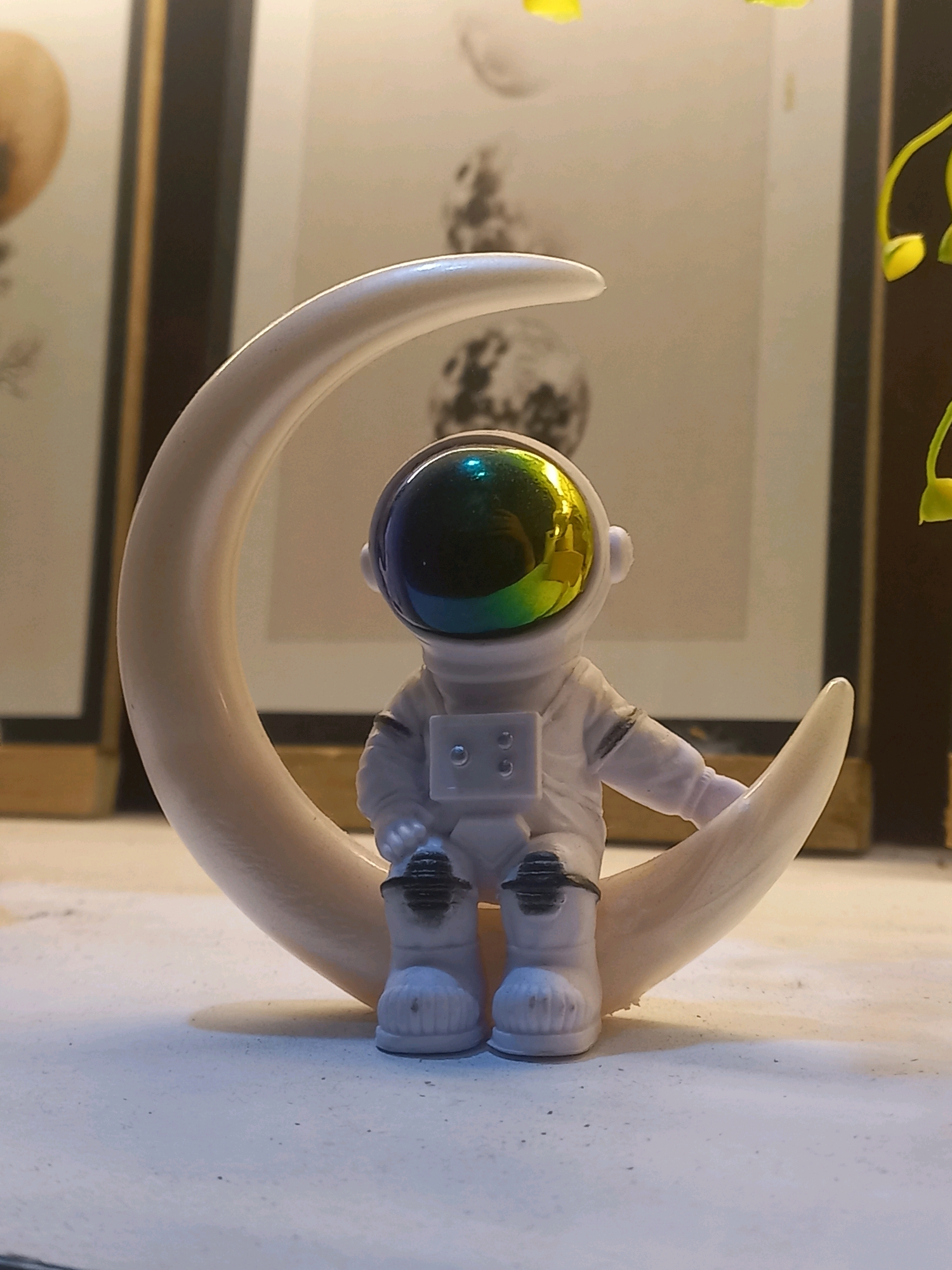 An astronaut figurine is seated on a crescent moon. The figurine is white with black accents on its boots and a reflective, iridescent visor. The moon is a pale, creamy color and curves around the astronaut. The background shows framed artwork with circular motifs, possibly depicting planets or celestial bodies, and a hint of greenery from a plant on the right. The lighting suggests an indoor setting, possibly a shelf or table. The image is a close-up shot focusing on the figurine and moon.