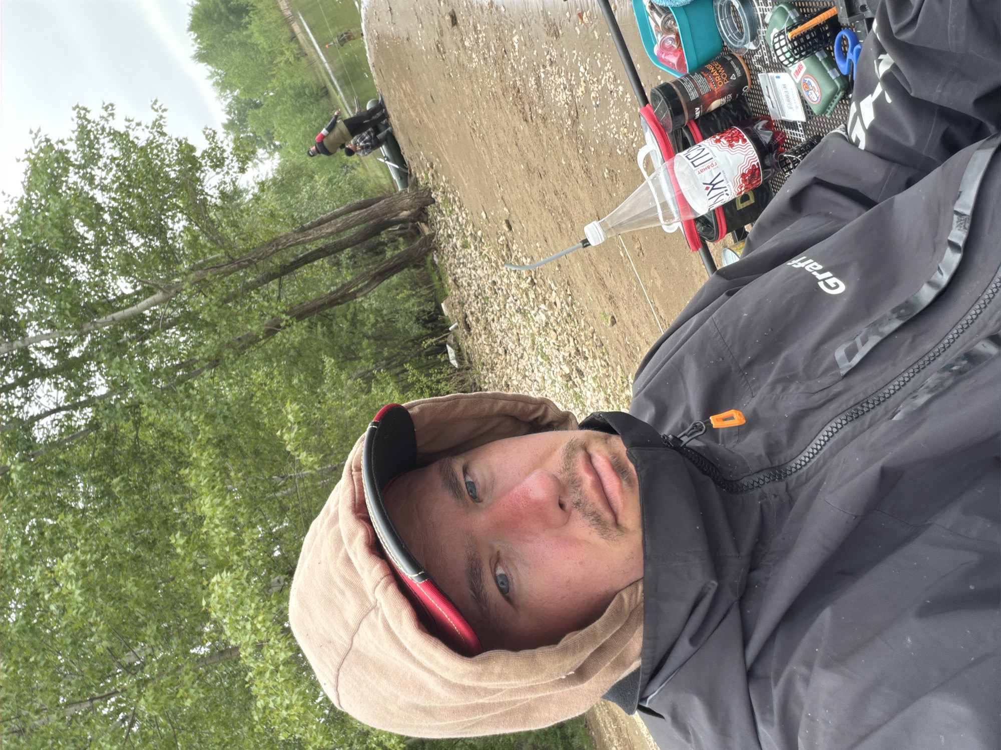 The image captures a close-up, slightly tilted portrait of a man, likely engaged in an outdoor activity, possibly fishing, by a body of water in Chaikovsky, Russia.
The main subject is a man in the foreground, looking directly at the camera. He has light-colored eyes, a light mustache, and a slightly reddish nose. He is wearing a black, possibly waterproof, jacket with the word "Graff" visible in white on the sleeve. Over his head, he has a tan-colored hooded sweatshirt or hood, and a red and black baseball cap is visible underneath the hood.
Behind the man, the scene extends to a natural environment. A dirt and gravel path or bank runs along what appears to be a river or lake. Lush green trees with light-colored bark are prominent on the left side of the frame, suggesting a wooded or riparian area. The sky above is overcast and light grey, indicating either a cloudy day or early morning/late afternoon light.
In the middle ground, another person is visible further down the bank, standing near what might be a vehicle or more fishing gear, suggesting companionship or another participant in the activity.
Several objects are clustered to the man's right, appearing to be fishing equipment. These include a blue container or bag, spools of fishing line, a small green box with the word "Rapala" visible on it, and a pair of blue scissors. There are also two cylindrical items: one black container with "AKWA" and "ORANGE" text, and another bottle or can, possibly a beverage, with "ГРАНАТ" (Pomegranate) written in Cyrillic. A clear plastic bottle with a dispenser nozzle is also present, likely for bait or attractant.
The overall impression is that of a person taking a break during a fishing outing on a mild, overcast day in a natural setting in Chaikovsky, Russia. The man's expression is neutral, and the focus is on his presence within this outdoor recreational context.
