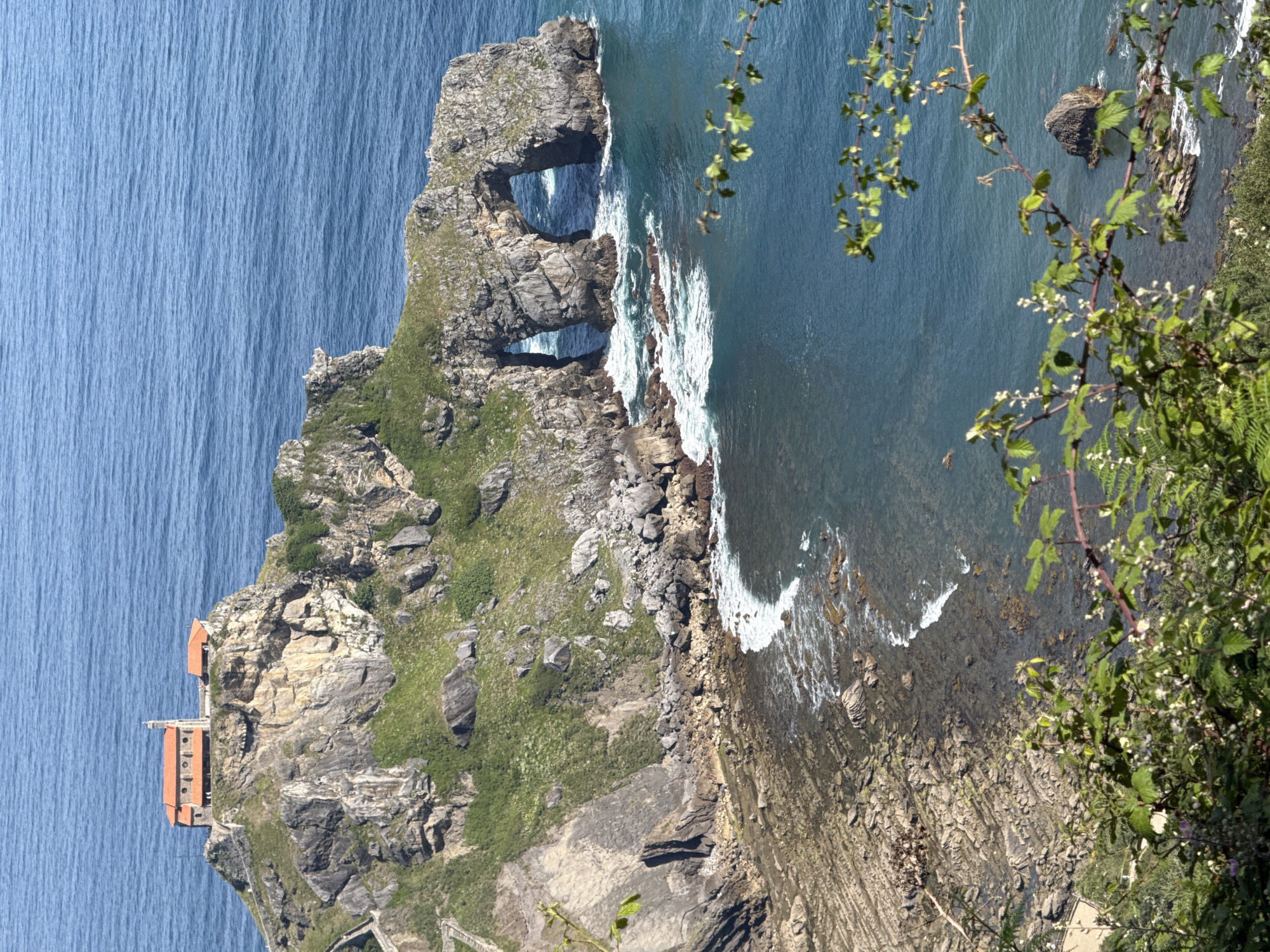 This is a vertical, eye-level, medium shot of Gaztelugatxe, a hermitage located on a small islet in the Bay of Biscay, connected to the coast of Bermeo, Spain by a man-made bridge. A stone church with a red tile roof sits atop the rocky outcrop, with a smaller building attached to its side. The island is rugged and covered in patches of green grass and scrub. The sea is a deep blue, with white waves crashing against the rocky shore and a prominent natural rock archway. In the foreground, out-of-focus green branches and leaves frame the scene, with a single dark rock visible in the blue water to the right. The day is sunny, with clear blue skies.