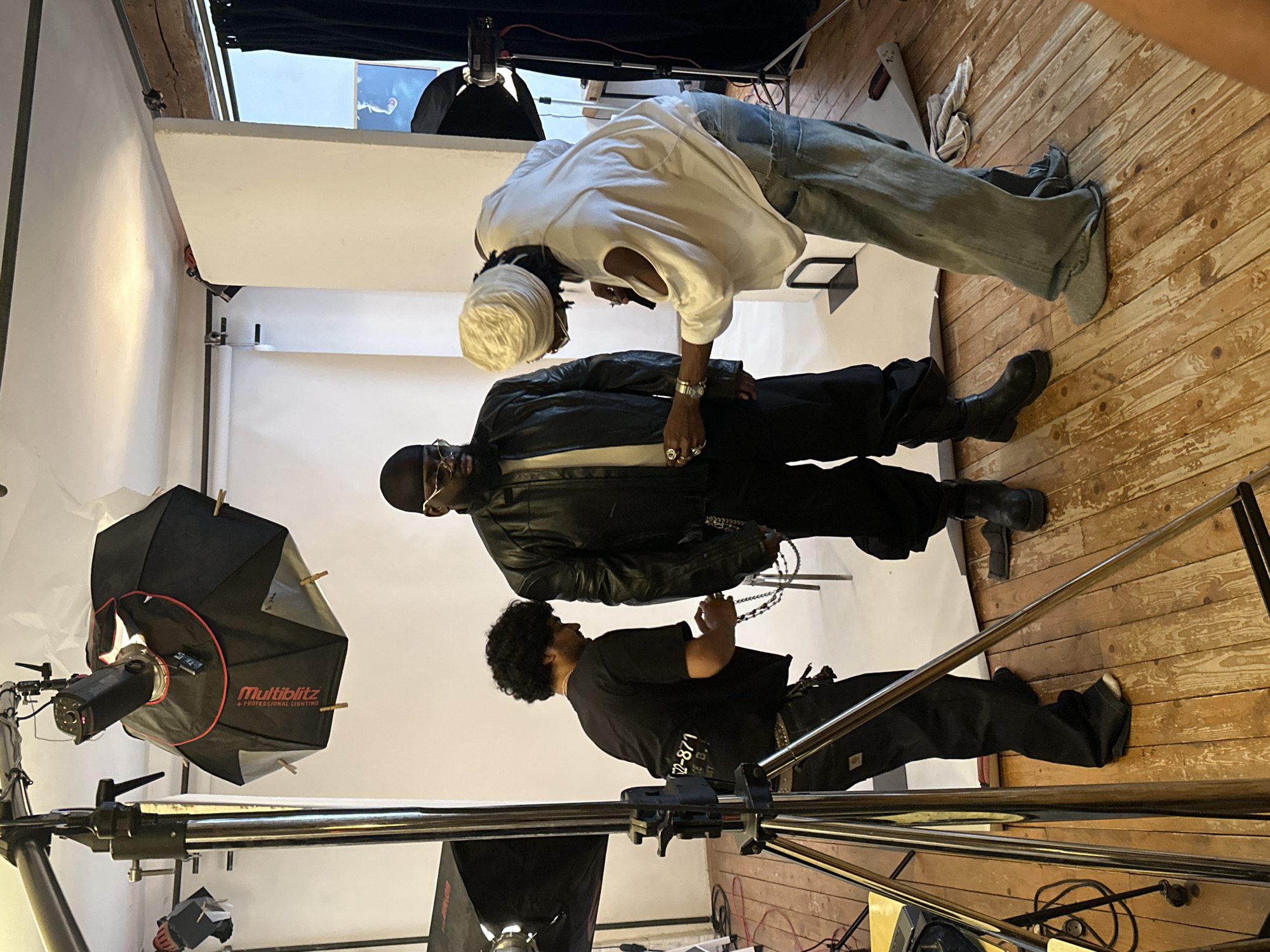 The image depicts three individuals in what appears to be a photography studio. The studio has a white backdrop, wooden flooring, and various pieces of photography equipment, including softboxes on stands. One person is wearing a white shirt, light-colored jeans, and a head covering. Another is dressed in a black leather jacket and black pants. A third person is wearing a black t-shirt and black pants. The three people are positioned close together, facing towards one another. The location of the city of Paris, France is unknown.