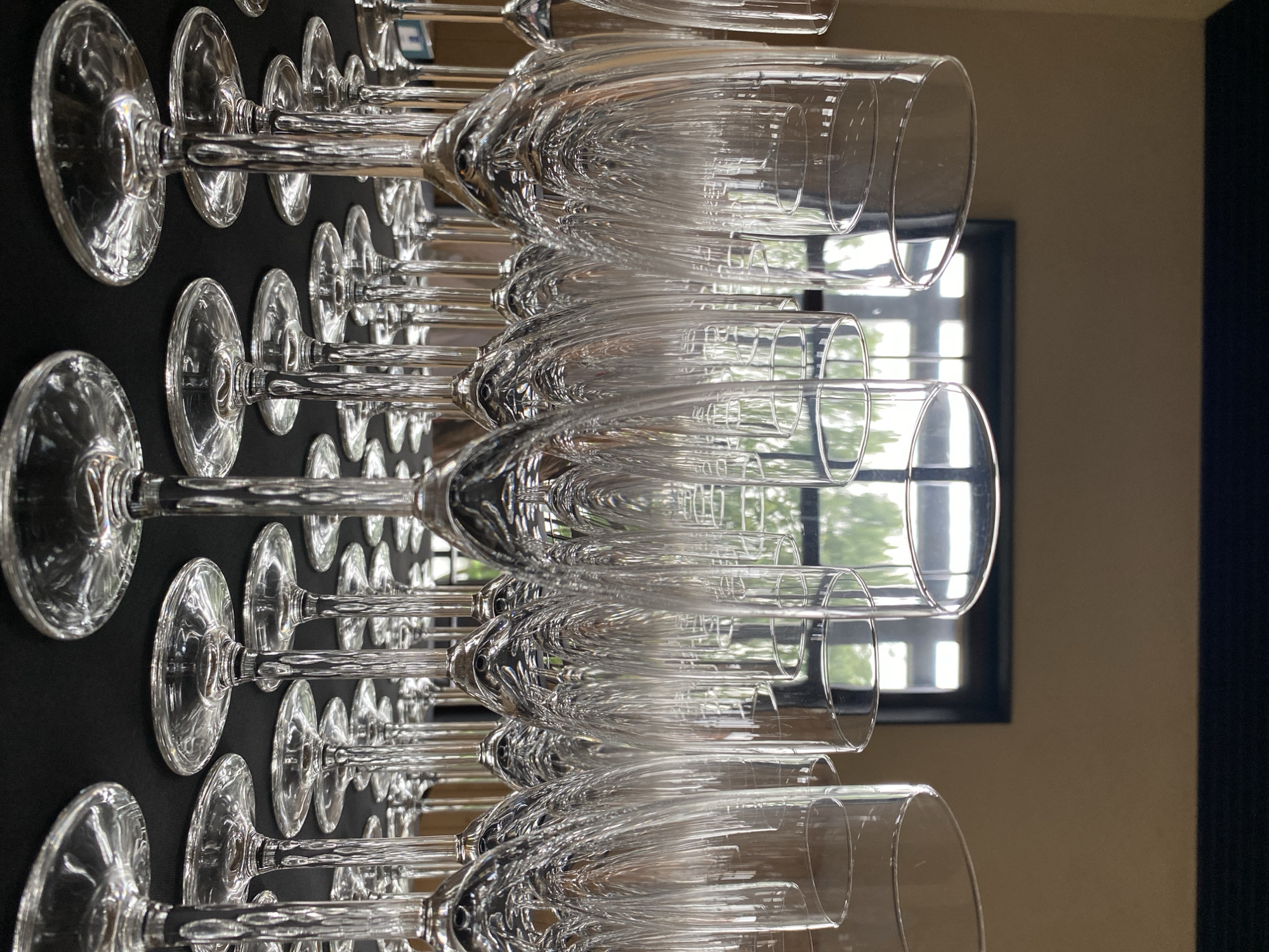 The image shows a close-up, low-angle view of many empty champagne flutes arranged on a black tablecloth. The flutes are clear glass and are set up in rows, with some slightly overlapping. The background is out of focus and appears to be a room with a window showing trees and sunlight. The scene is indoors, but the presence of greenery visible through the window suggests it might be in a location with access to the outdoors, such as a restaurant, event venue, or a private residence. There are no people or visible actions occurring in the image. The image does not provide any information or context to determine its location in the city of Buk, Poland.