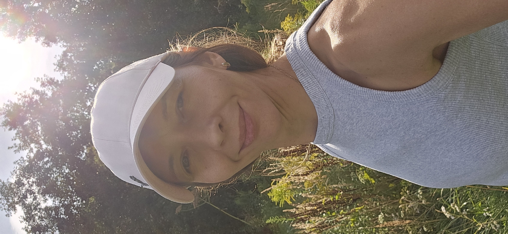 The image shows a close-up selfie of a woman with light brown hair, wearing a white baseball cap and a gray tank top. She is outdoors, with a blurred background of trees and vegetation, suggesting a natural or park-like setting. The lighting indicates it is daytime, with sunlight creating a lens flare in the upper right portion of the image. Her expression is a slight smile. The location is described as Мазалаўскі сельскі Савет, Belarus.