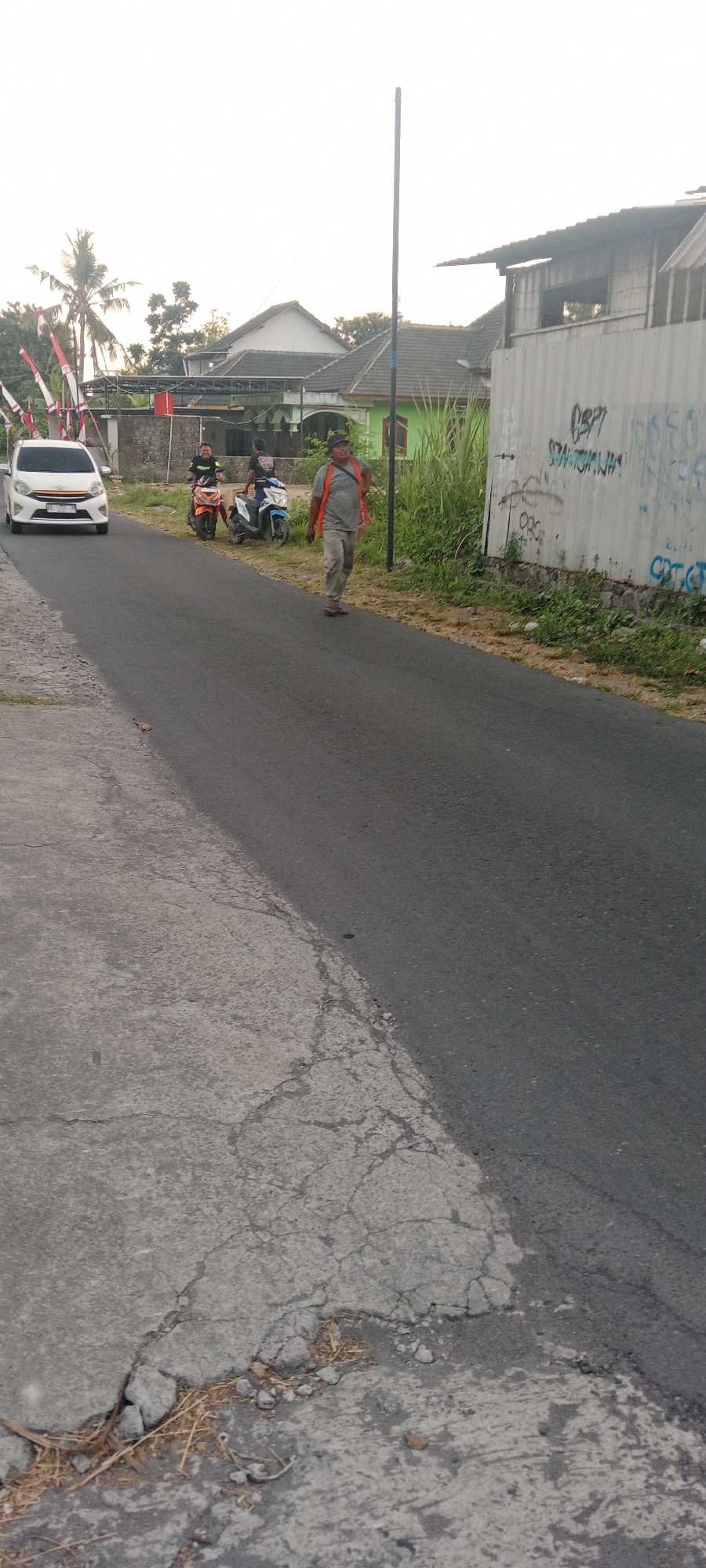 The image depicts a street scene in Maguwoharjo, Indonesia, under an overcast sky. On the left side of the asphalt road, a white car is driving towards the viewer. Further down the road, two individuals are seated on motorcycles, one orange and one blue. To the right of the motorcycles, a person wearing a grey shirt, grey pants, a cap, and an orange vest is walking away from the viewer. Behind the walking person, a tall, thin pole stands vertically. The background features houses with tiled roofs, some with green walls, and lush vegetation. To the far right, a corrugated metal wall is covered in graffiti. The roadside on the left is a cracked concrete surface with dry grass and debris.