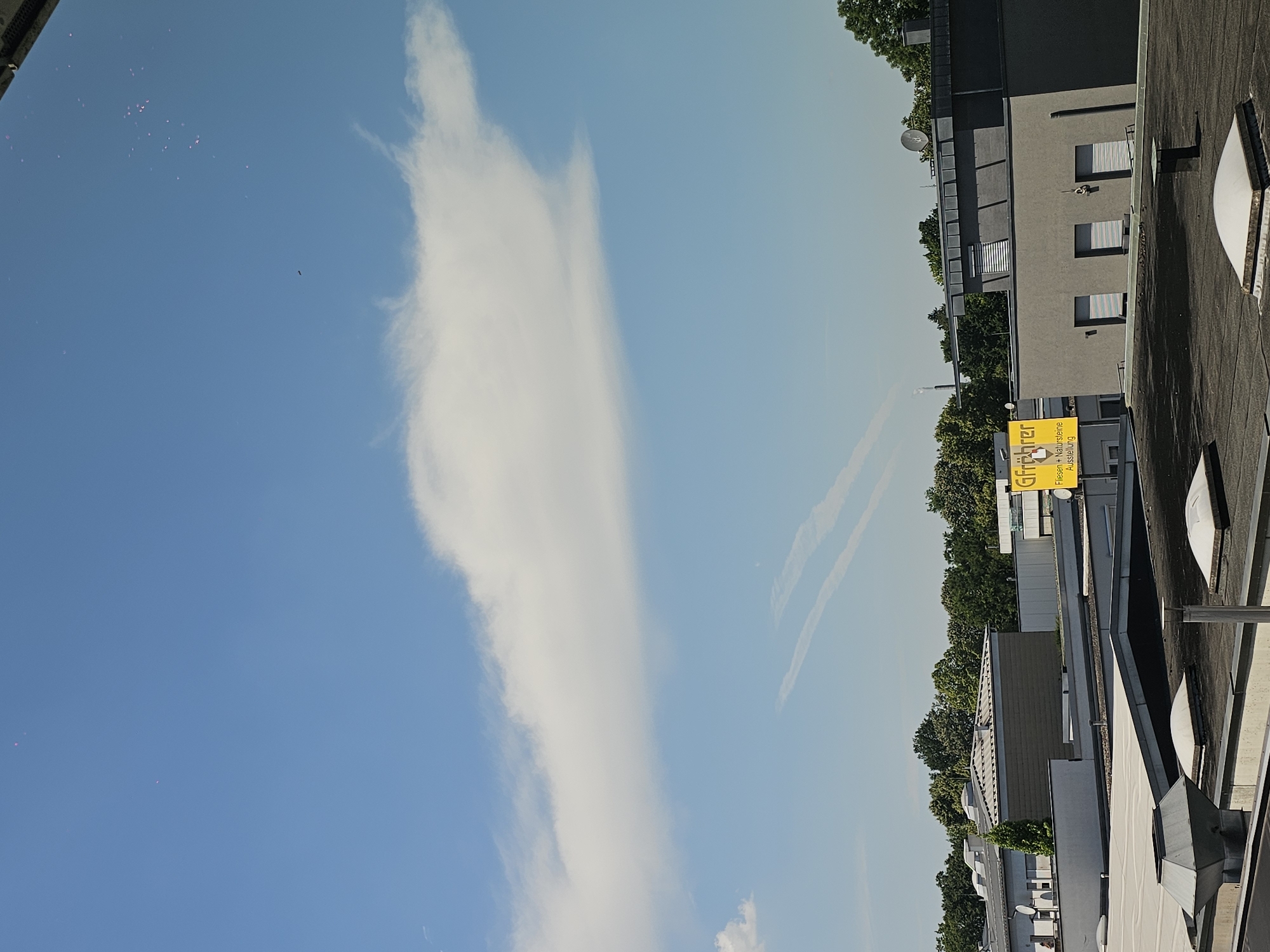 The image depicts an outdoor scene, likely from a rooftop, showcasing a bright blue sky with a large, elongated white cloud stretching across a significant portion of the frame. Below the sky, a line of trees and modern buildings can be seen. One building has a dark gray facade with visible windows and shutters, and another section of the building appears lighter gray. A prominent yellow sign with black text is affixed to the side of a building, partially obscured by a tree. The sign reads "Grührer" and below that, "Fliesen + Natursteine Ausstellung". There are also several rooftop structures visible, including vents and a satellite dish. In the upper left portion of the sky, a cluster of small, pinkish dots are scattered, possibly balloons or reflections. There are also faint, parallel white lines in the sky, suggesting contrails from an aircraft. The overall impression is of a clear day with a few scattered clouds, likely in the afternoon due to the bright sunlight. The scene appears to be urban or suburban, with a mix of natural elements and man-made structures.