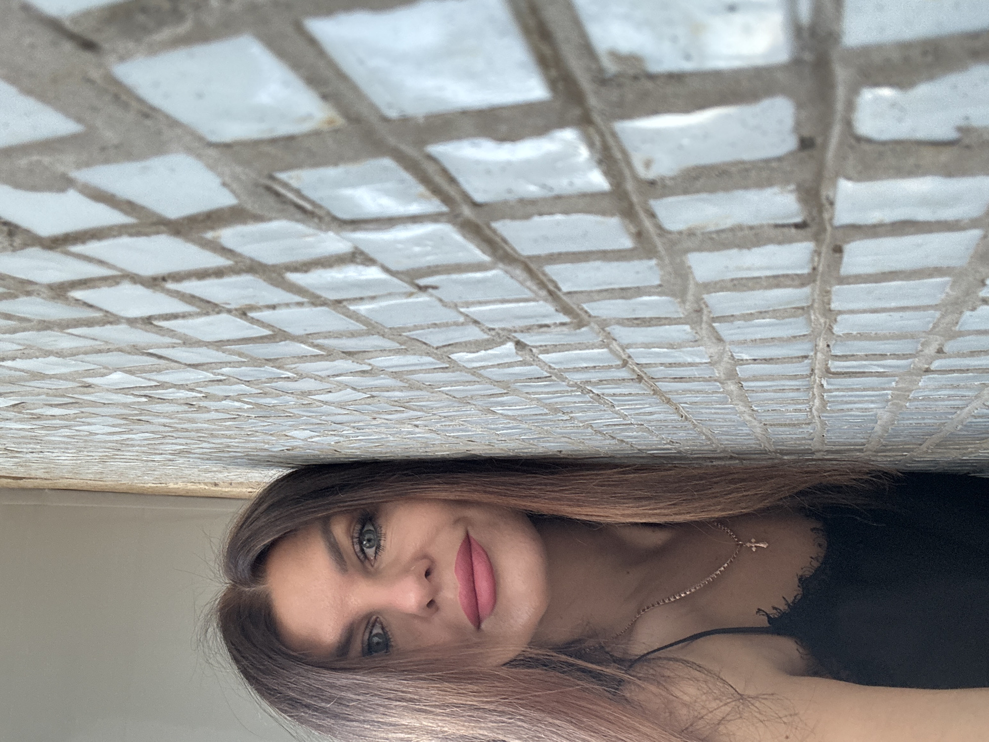 The image shows a woman posing against a tiled wall. The tiles are small and square, with a light color. The woman has shoulder-length hair, light eyes, and a necklace with a cross pendant. The scene is likely indoors. There is no discernible information to link this to the city of Chaikovsky, Russia.