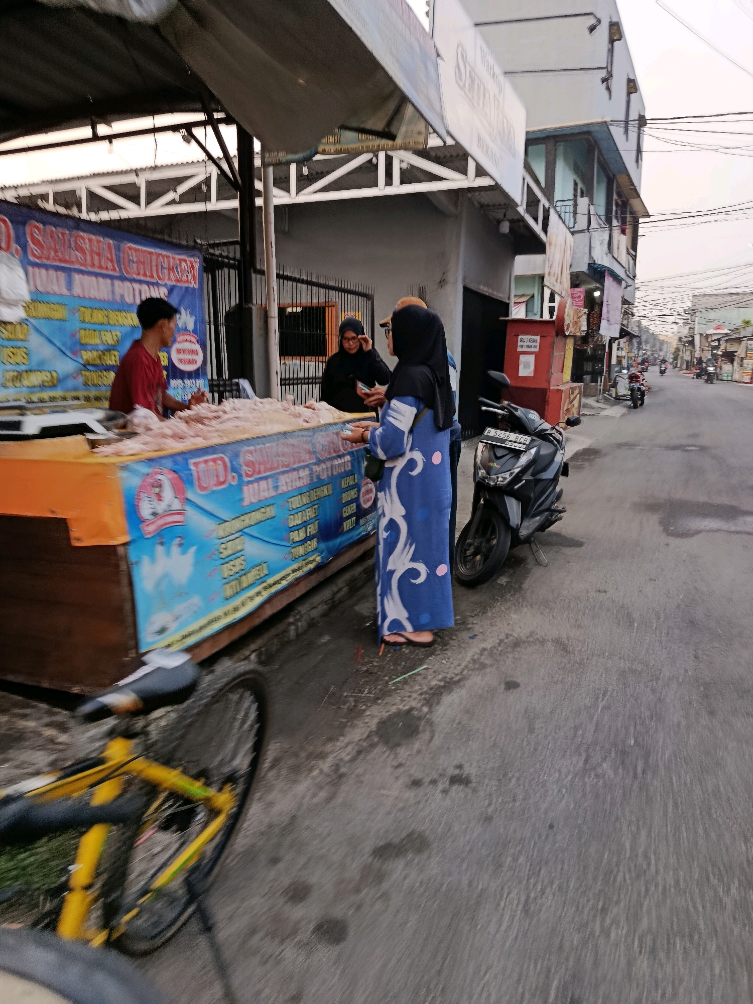 The image depicts an outdoor scene, likely a street market, in Bekasi, Indonesia. In the foreground, a yellow bicycle is partially visible on the left side. To the right, a stall sells chicken products, indicated by a blue banner with "UD. SALSHA CHICKEN" and "JUAL AYAM POTONG" (selling cut chicken). A man in a red shirt stands behind the stall, attending to the display of raw chicken. Two women are interacting at the stall; one, wearing a black hijab, appears to be purchasing chicken, while another in a blue patterned dress stands next to her. A black motorcycle is parked on the right side of the street. The background shows buildings, including a multi-story structure with balconies, and a street lined with other stalls and people. The overall environment suggests a bustling urban commercial area.